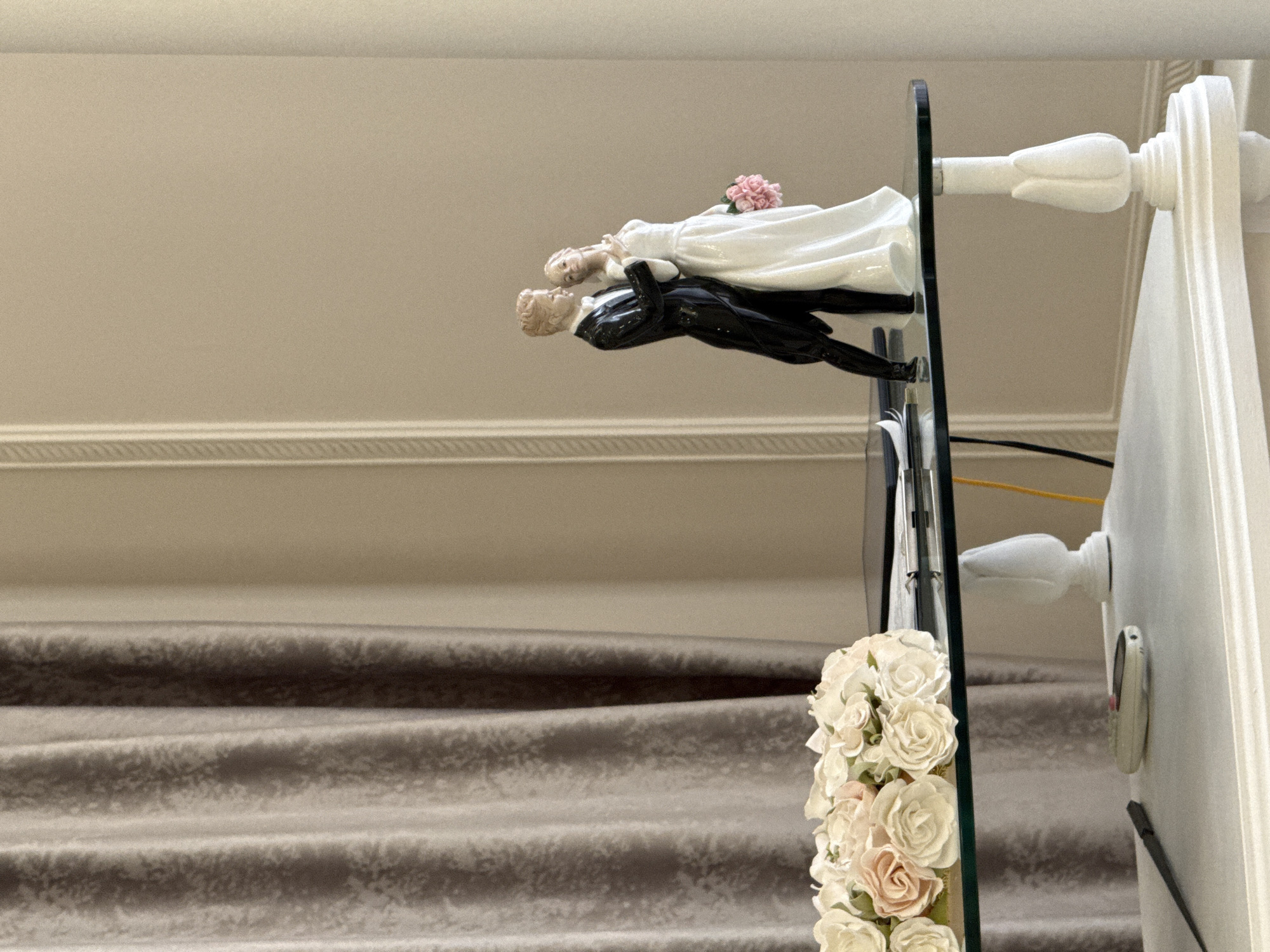 The image shows a wedding cake topper of a bride and groom standing on a glass table, with a bouquet of artificial roses in soft pink and cream colors to the left. The cake topper depicts a groom in a black tuxedo and a bride in a white wedding dress, holding a pink bouquet. Behind the table, a beige wall with decorative molding is visible, along with draped grey curtains. A tablet or e-reader and some cables are on the glass table. Below the glass table, a white table surface holds a black stylus and a cream-colored remote control. The scene appears to be indoors, in a room with muted lighting.