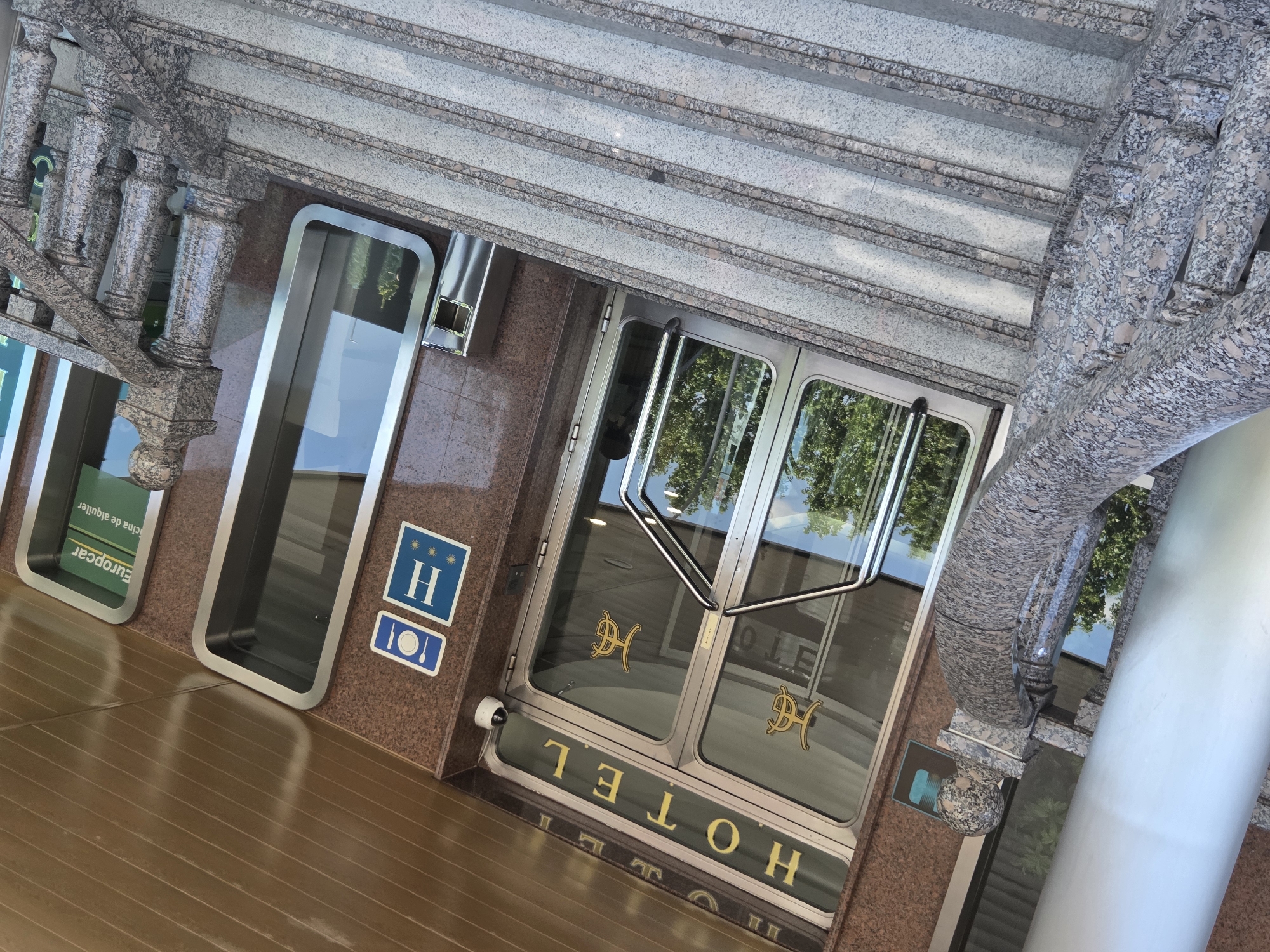 The image shows the entrance to a hotel and a Europcar rental agency. The hotel entrance features double glass doors with the word "HOTEL" in gold lettering above them. Two hotel logos, appearing to be stylized "HG" or "GH", are visible on the glass doors. Below the "HOTEL" sign, there are two blue and white signs: one with a fork and knife icon and a "H" with three stars, indicating a hotel. Adjacent to the hotel entrance, to the right, is a Europcar rental agency with a green sign featuring the Europcar logo and text in Spanish that translates to "rental office". The entrance is framed by granite-clad walls and a polished granite balustrade and staircase. The scene is illuminated by natural daylight. A silver trash receptacle is located to the right of the hotel entrance. The location is identified as La Estación, Spain.