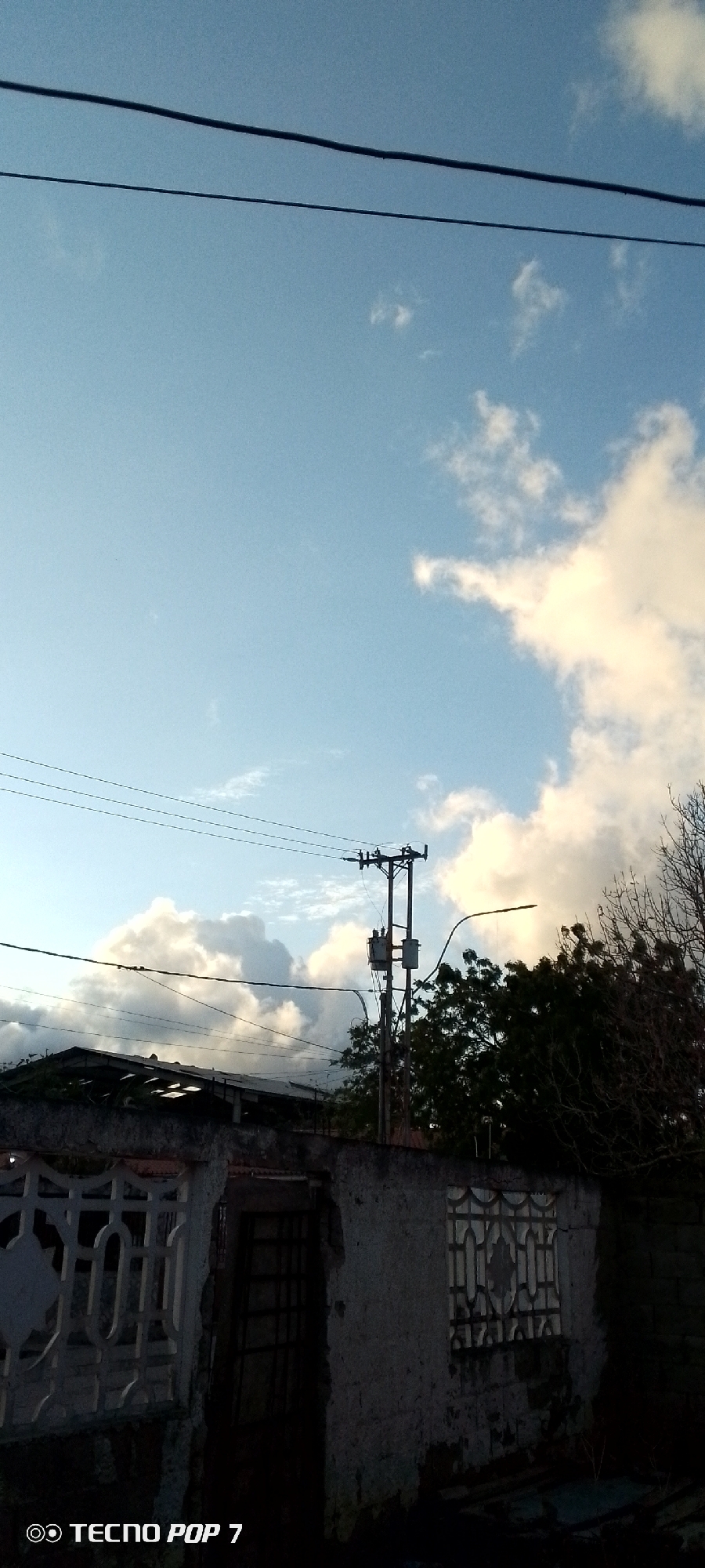 The image shows a section of Altagracia, Venezuela. The sky is blue with scattered white clouds. Electrical wires stretch across the top portion of the frame. Below, a utility pole with transformers is visible, partially obscured by a tree. In the foreground, a weathered wall or fence is constructed from concrete or stone. The wall contains decorative patterns and a gated opening. The "Tecno Pop 7" watermark is visible in the lower left corner.