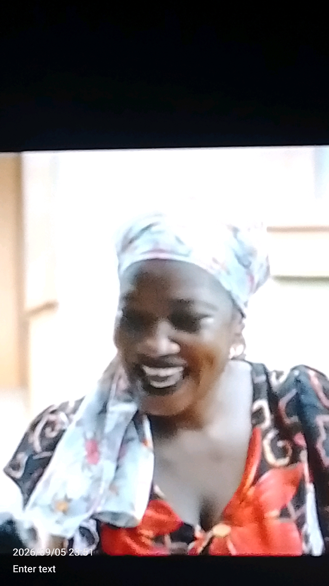 A woman with dark skin is shown from the chest up, smiling widely. She is wearing a headscarf with a floral pattern in light blue, pink, and white. She also wears a colorful dress with large red and black floral designs. The background is out of focus and appears to be indoors, with warm-toned walls. Text overlays at the bottom of the image read "2026/09/05 23:51" and "Enter text". This image is not geographically specific to Lokoja, Nigeria, and no details within the image can be used to confirm or deny this location.
