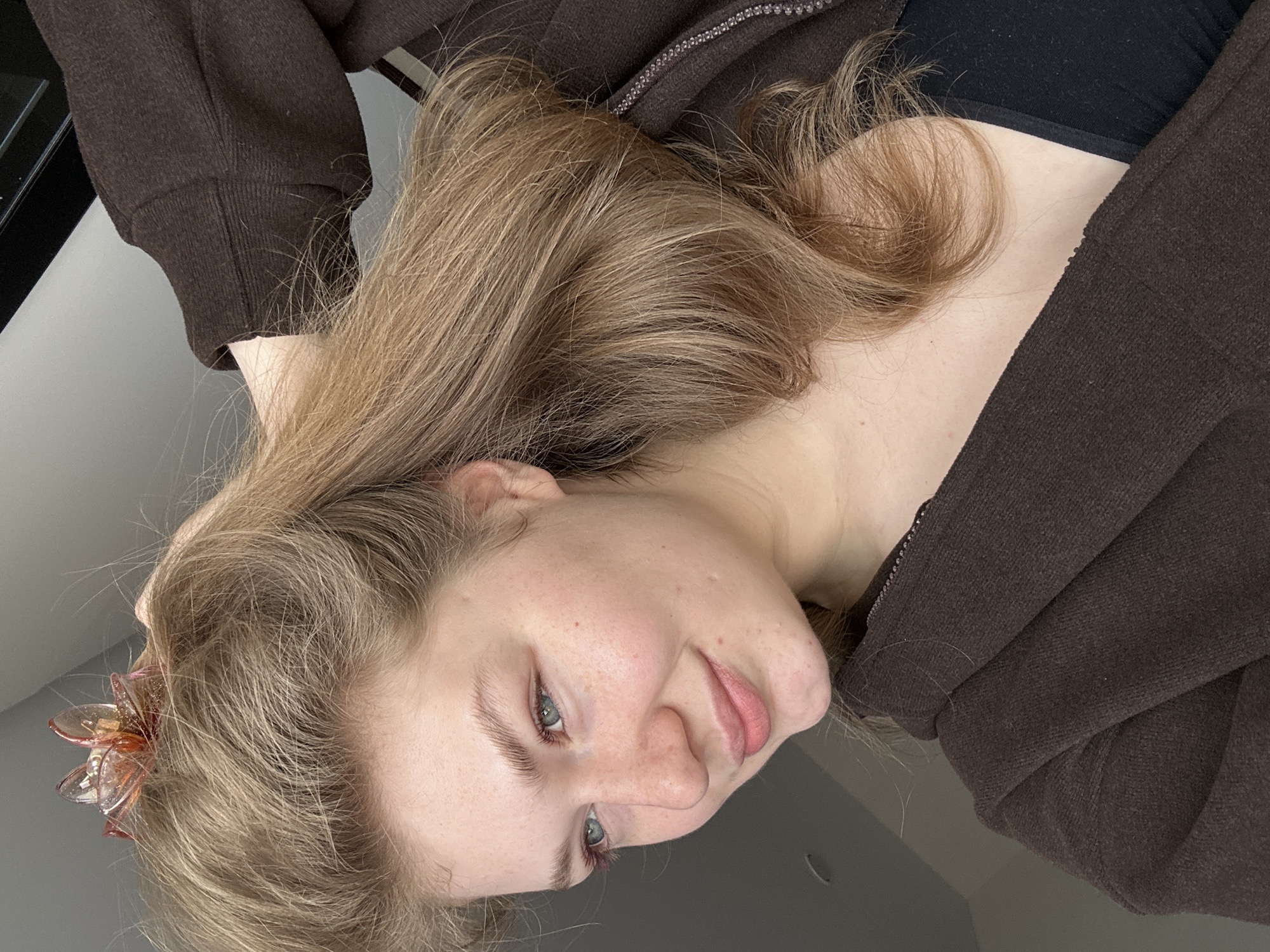 A young fair-skinned individual with light blonde hair is seen in a close-up shot. They are wearing a dark brown zip-up hoodie over a black top. A decorative flower hair clip is visible in their hair. The background appears to be a plain light gray surface. There is no evidence in the image to indicate the location is in Kazan, Russia.
