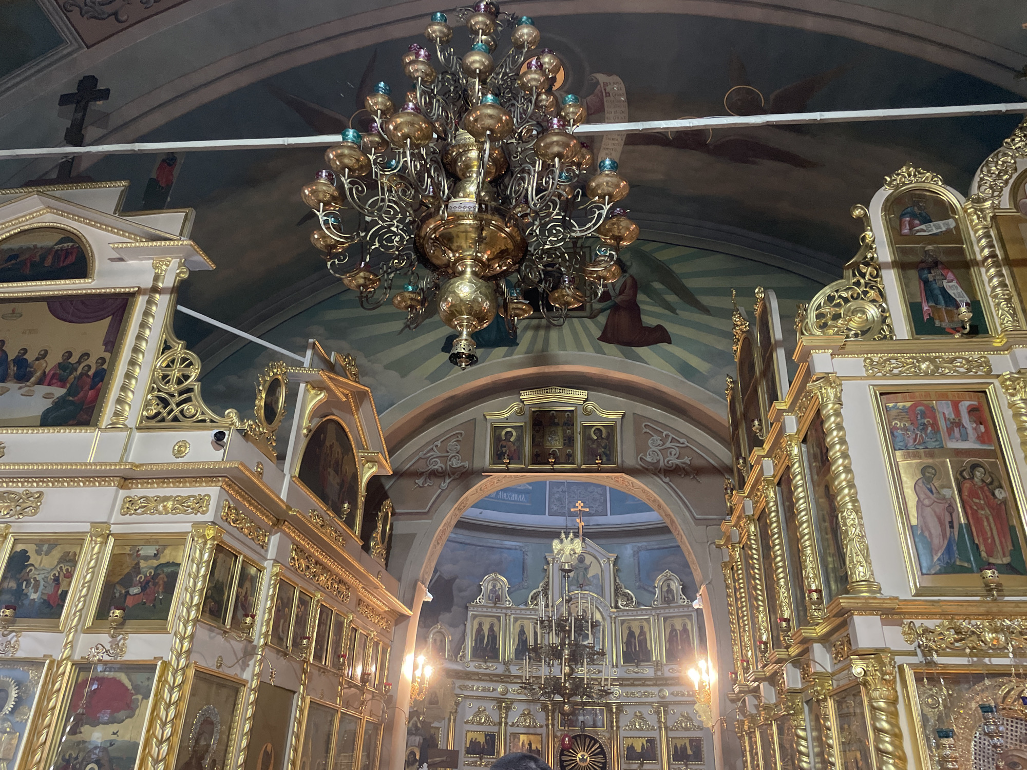 The image shows the interior of a Russian Orthodox church, likely in Ivanisovo, Russia. A large, ornate chandelier hangs from the ceiling. The walls are adorned with gold-framed religious icons and paintings. The ceiling features religious artwork and a black cross. The architectural design is characterized by arches and golden detailing. The overall environment suggests a space of worship and historical significance.