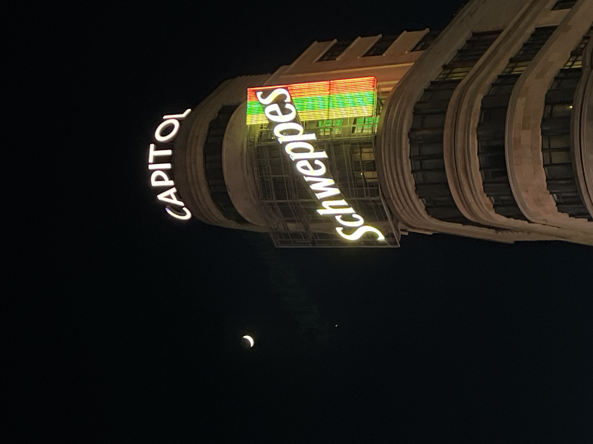 The image shows the Capitol building in Madrid, Spain, at night. The building's facade is visible, with the illuminated "CAPITOL" and "Schweppes" signs prominently displayed. A crescent moon and a distant point of light, possibly a star or planet, are visible in the dark sky below the building. The building's architecture is characterized by a series of rounded, stacked layers. A section with colorful lights sits atop a portion of the building.