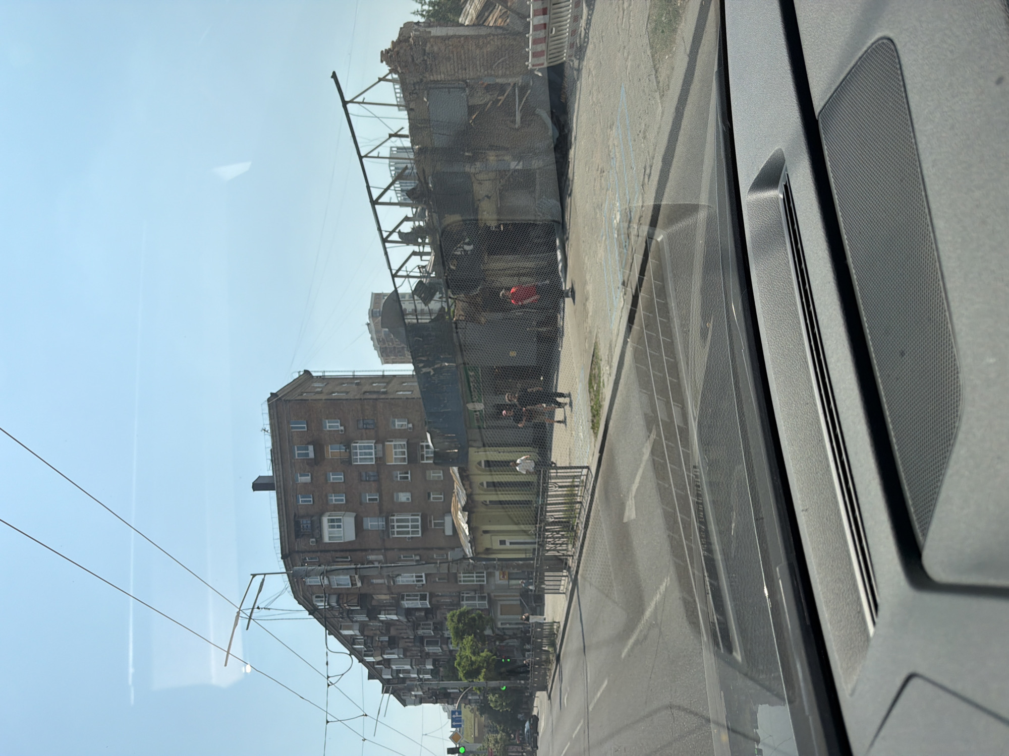 In Kyiv, Ukraine, a wide street in a city environment is depicted on a sunny day. On the right side of the street, a building appears to have been destroyed, with its roof and some walls missing, exposing a skeletal metal structure. Debris and construction barriers are visible near the damaged structure. Across the street, a multi-story brick apartment building with numerous windows and balconies stands intact. Several people are seen walking on the sidewalk in front of the apartment building, and one person in a red shirt stands near the damaged building. The street is paved and marked with lane lines, and traffic lights are visible in the distance. The scene is captured from the perspective of someone inside a vehicle, as evidenced by the car's dashboard and windshield in the foreground.