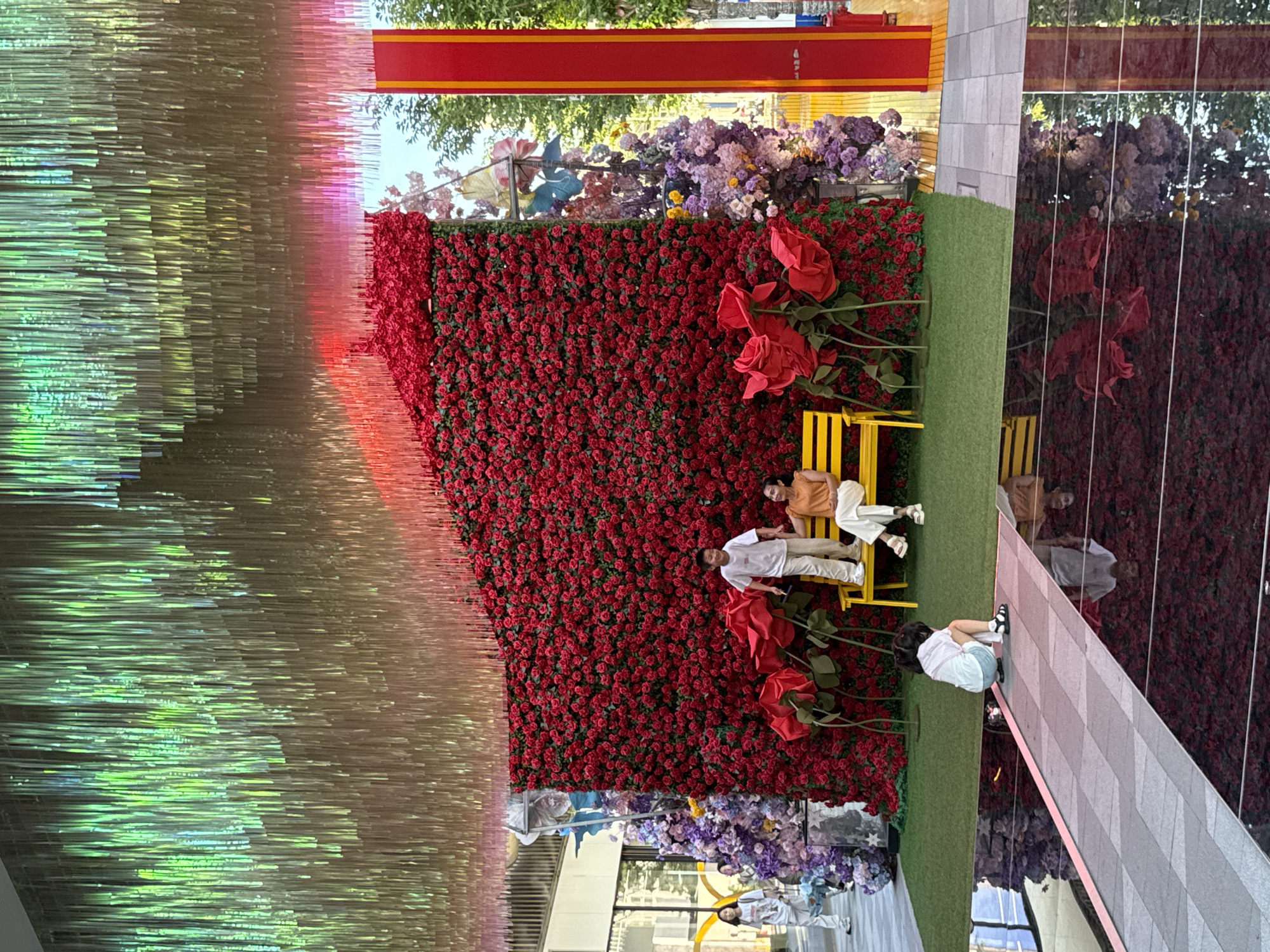 An outdoor scene in Yiwu, China features a vibrant floral display with a wall of red roses as the centerpiece. A yellow bench is placed in front of the rose wall, occupied by a woman in an orange top and white pants, and a boy in a white t-shirt and beige pants. The boy is standing and holding the woman's hand. To the left of the rose wall, a young child wearing a light-colored t-shirt and blue shorts is crouching on a walkway. The walkway has a reflective surface, mirroring the surroundings. Hanging above the display are iridescent streamers in shades of green, gold, and pink. To the right of the rose wall, there are tall, purple and pink floral arrangements and a red and yellow vertical structure. In the background, trees and buildings are visible.