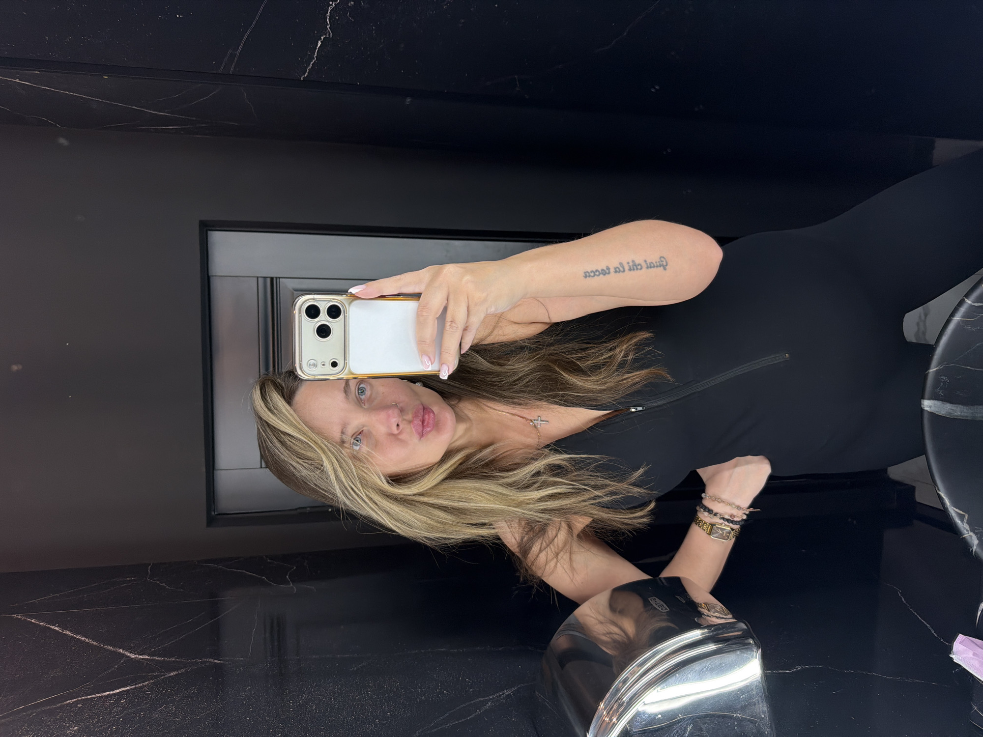 A woman with long, blonde hair is taking a selfie in a bathroom. She is wearing a black, zippered jumpsuit and several bracelets. A cross necklace is visible around her neck. She has a tattoo on her left forearm. The bathroom has dark, marbled walls and a silver hand dryer is visible on the left. The woman's facial expression is one of puckering her lips.