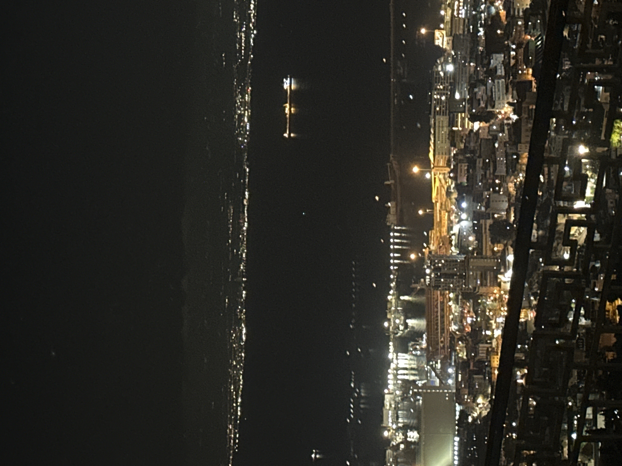 This is a nighttime aerial shot of Haifa, Israel, looking out over the bay. The foreground features a blurred railing, suggesting the photo was taken from a high vantage point, possibly a building or hill overlooking the city. Below this, the city lights of Haifa twinkle, creating a dense urban landscape. Further out, the dark waters of Haifa Bay are visible, dotted with various lights from boats and possibly offshore installations. A prominent cargo ship is illuminated in the bay, likely at anchor or navigating. In the distance, the opposite shore of the bay is lined with more city lights, extending into the horizon. The sky is dark, indicating it is well after sunset.