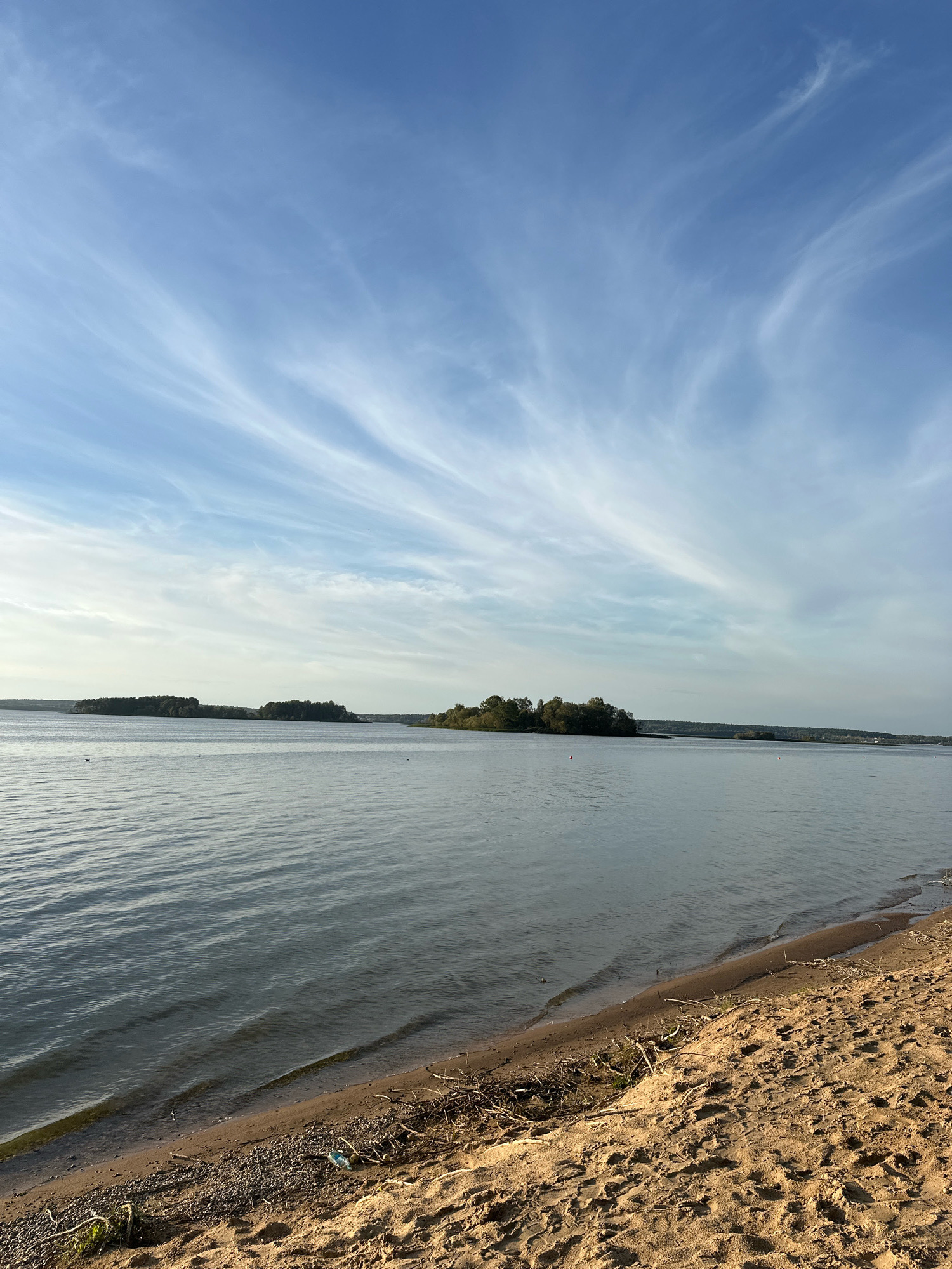 The image depicts a serene natural landscape, likely a reservoir or large body of water, with a sandy shore in the foreground. The water is calm, reflecting the blue sky and wispy white clouds. On the horizon, several densely wooded islands dot the water, adding depth to the scene. The shoreline is composed of sand interspersed with small pebbles and scattered driftwood. A small piece of blue plastic debris is visible on the sand near the water's edge. The overall impression is one of peacefulness and natural beauty, possibly a public recreation area.