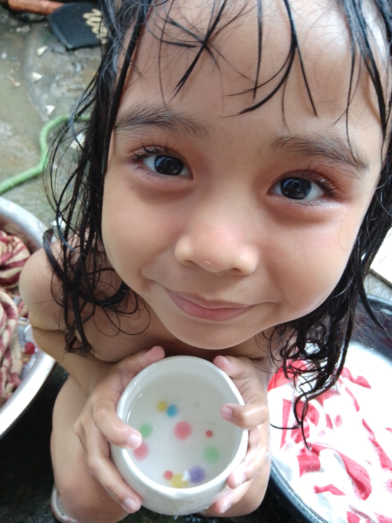 A close-up photograph captures a young child, appearing to be female and of Asian descent, approximately 3-6 years old, engaged in water play outdoors in Davao City, Philippines. The child's dark hair is wet and matted, with strands falling across her forehead and along her face. She is positioned below the camera's perspective, looking upwards and directly into the lens with wide, dark eyes and a distinct smile.
She holds a small, white bowl with both hands, positioned at approximately chest level. The bowl contains clear water with multiple small, colorful, spherical objects, including pink, light blue, yellow, purple, and green beads, likely water beads or small marbles.
The background reveals an outdoor utility area with a wet, dark-colored ground, consistent with concrete or pavement, featuring scattered small debris. To the left, the edge of a metallic basin or tub is visible, containing patterned fabric (red and off-white). A green garden hose lies on the ground. To the right, another dark basin or tub holds more red and white patterned fabric. The scene is illuminated by natural daylight.