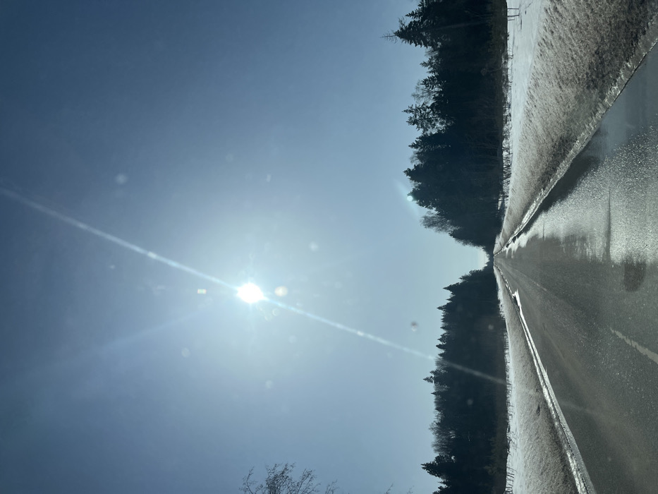 The image depicts a bright, rural road scene, reportedly located near the settlement of Нарынка, Russia. The sky is clear and blue, dominated by a glaring sun in the upper-left quadrant, which produces a prominent sunburst and lens flare across the upper portion of the frame. A straight, two-lane asphalt road extends into the distance, appearing wet with melted snow and puddles, and reflecting the sunlight. Elevated banks of dirty, melting snow line both sides of the road. Beyond the snowbanks, dense forests, primarily composed of dark evergreen trees interspersed with some bare deciduous trees, flank the road to the horizon. No individuals or other vehicles are visible. The perspective suggests the image was captured from inside a moving vehicle.