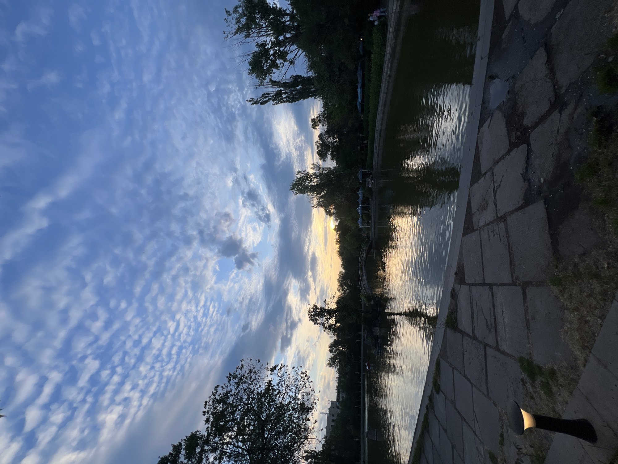 The image depicts a serene waterscape at dusk, likely in a park setting, given the presence of trees and a paved walkway bordering a body of water. The sun is setting, casting a warm, golden glow on the horizon and reflecting on the rippling surface of the water. The sky is a canvas of soft blues and grays, with streaks of white clouds that appear to be illuminated by the fading sunlight.
The water's edge is lined with large, flat stone slabs, creating a pathway that leads towards the foreground. A low, dark-colored lamp with a warm light is visible on the pathway, suggesting it's an area designed for evening ambiance. Trees with dark green foliage line the opposite bank of the water, their silhouettes contrasting with the brighter sky. Some structures, possibly gazebos or covered seating areas, can be seen amidst the trees along the water's edge. In the distance, a few indistinct figures, likely people, are visible near the far bank, adding a sense of scale and quiet activity to the scene.
The overall mood is tranquil and contemplative, evoking a sense of peace and the beauty of nature at twilight. The light filtering through the clouds and reflecting on the water creates a painterly effect. The orientation of the image is such that the sky occupies the left half, and the water and pathway occupy the right, with the sunset occurring in the middle distance. There is no visible text in the image.