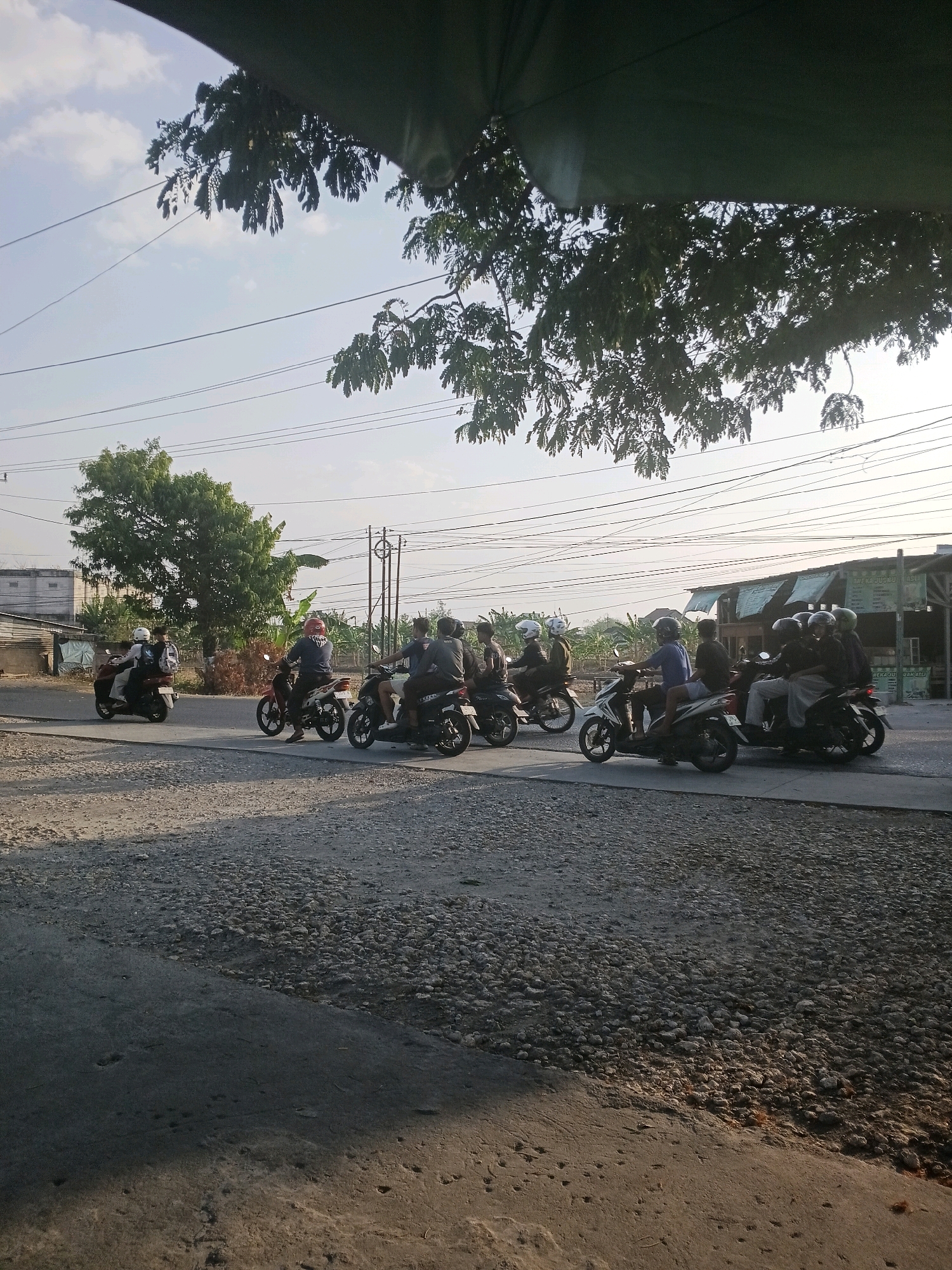 In Purwodadi, Indonesia, a group of people ride motorbikes in what appears to be a rural or suburban setting. The scene captures a moment on a road with a gravel shoulder and a paved surface. Several motorbikes, each carrying at least one passenger, are traveling in the same direction. The individuals are wearing helmets, and some have multiple people per motorbike. In the background, there are buildings, trees, and utility poles with numerous wires. The sky is partly cloudy, and the sunlight suggests it is either early morning or late afternoon. The overall impression is a common mode of transportation in the area.