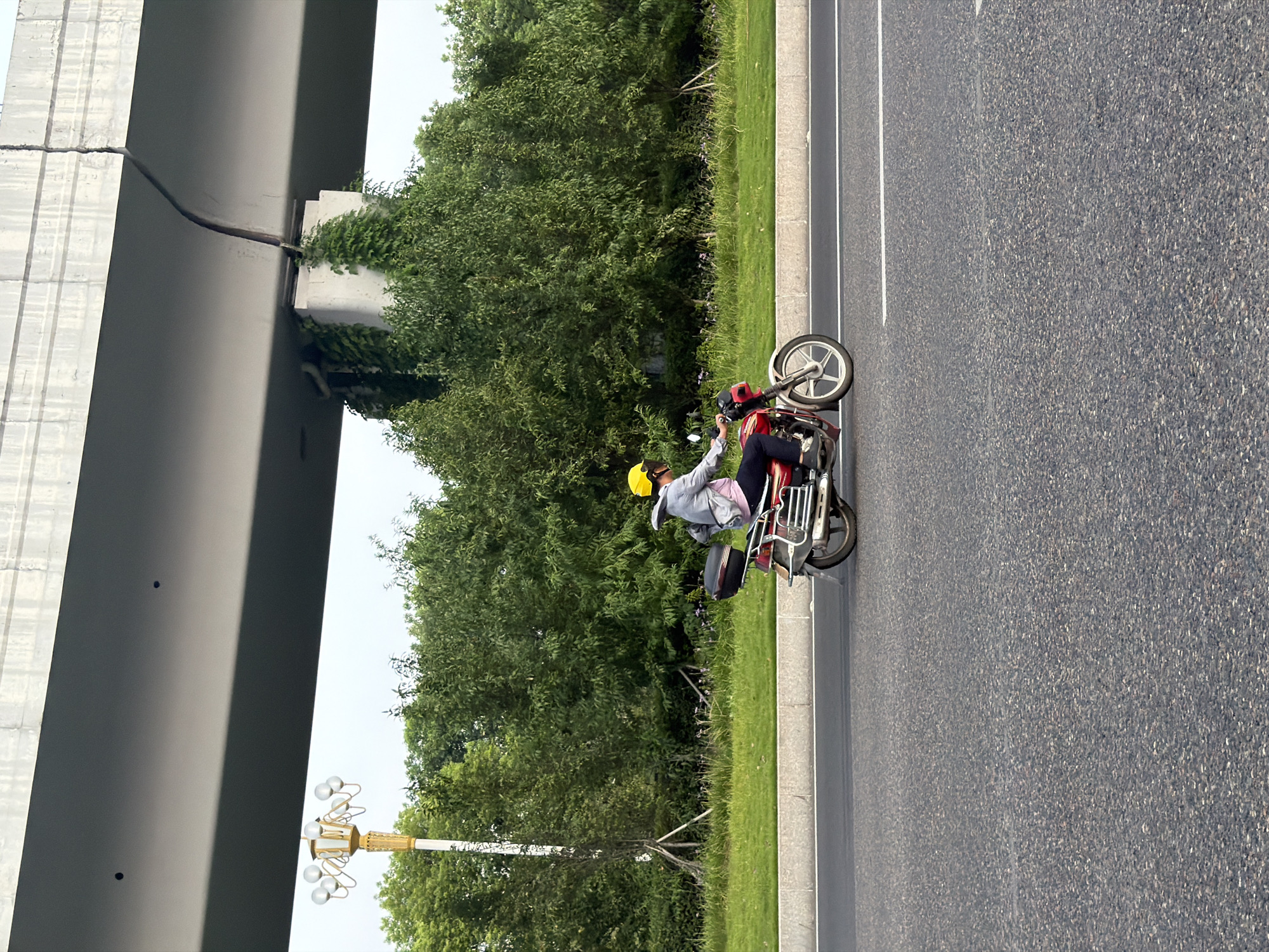 A person wearing a yellow helmet and a gray jacket is riding a red motorcycle on a paved road. The motorcycle has a black storage box on the back. The person is in the middle of the frame, moving from left to right. In the background, there is a lush green treeline and a raised concrete structure that appears to be an overpass or bridge. A street lamp with multiple globes is visible to the left of the motorcycle, and a white line marks the edge of the road. The overall scene suggests an urban or suburban environment, possibly in the city of Jiangjin, China, based on the provided context. The lighting indicates it is daytime, with diffused light suggesting an overcast or slightly hazy sky.