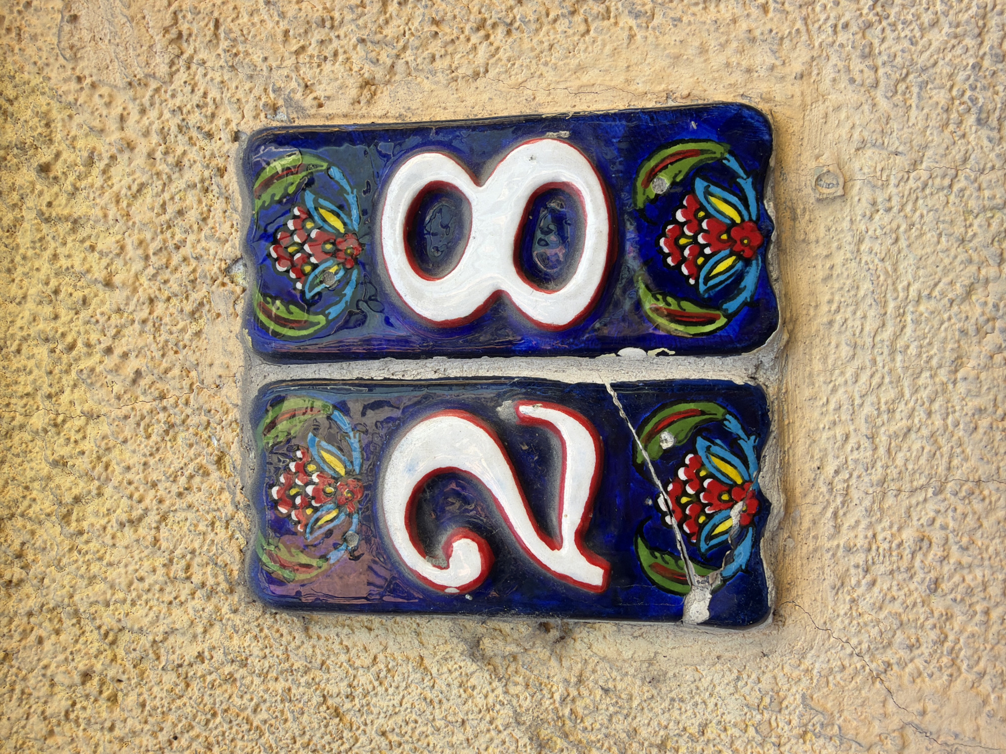 The image displays a ceramic house number plaque with the numerals "28" set against a textured, light brown wall. The plaque itself is rectangular with rounded corners and features a dark blue background decorated with colorful floral motifs. Each numeral, "2" and "8", is rendered in thick white paint with a red outline and is placed on its own decorative panel. The "2" is on the left and the "8" is on the right. Both panels are adorned with stylized red, yellow, and green flowers. The plaque is cracked in several places, indicating age or damage. The wall it is mounted on appears to be stucco or concrete, with visible surface imperfections and hairline cracks. The lighting suggests it is daytime, with soft shadows present. This plaque is located in Istanbul, Turkey, on a building wall. There are no people or animals visible in the image.