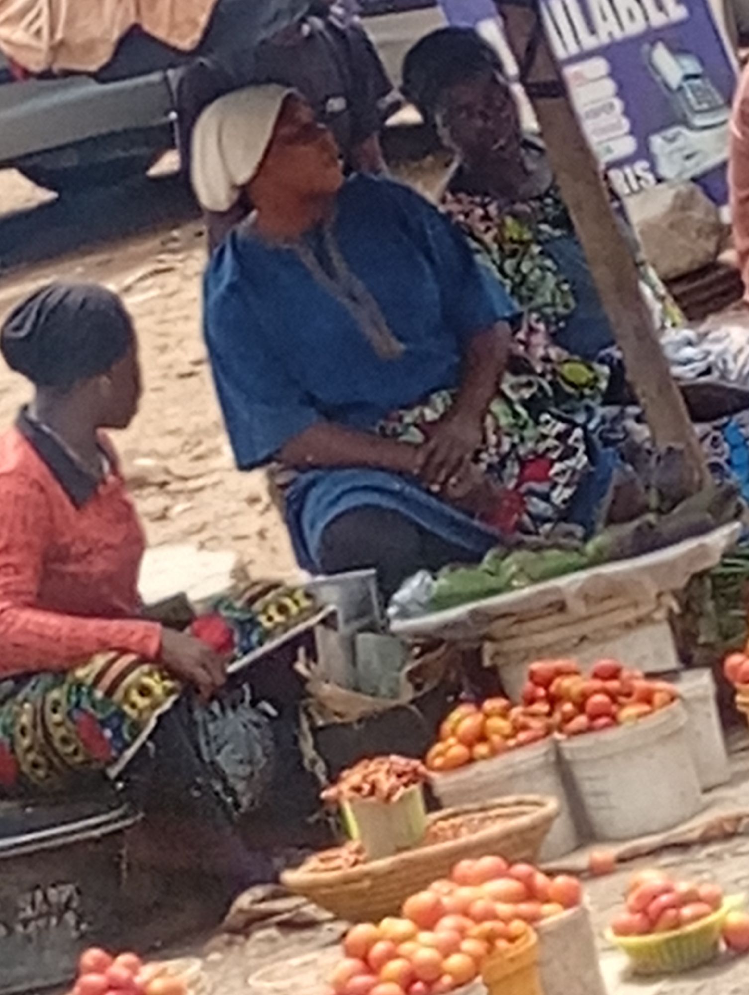 In Maiburiji, Nigeria, three women are selling goods at an outdoor market. One wears a white head covering and a blue dress; another wears a black head covering and a red top with patterned skirt. A third woman wears a floral top. Baskets and buckets of tomatoes are visible, along with other items for sale. The setting appears to be a busy marketplace.