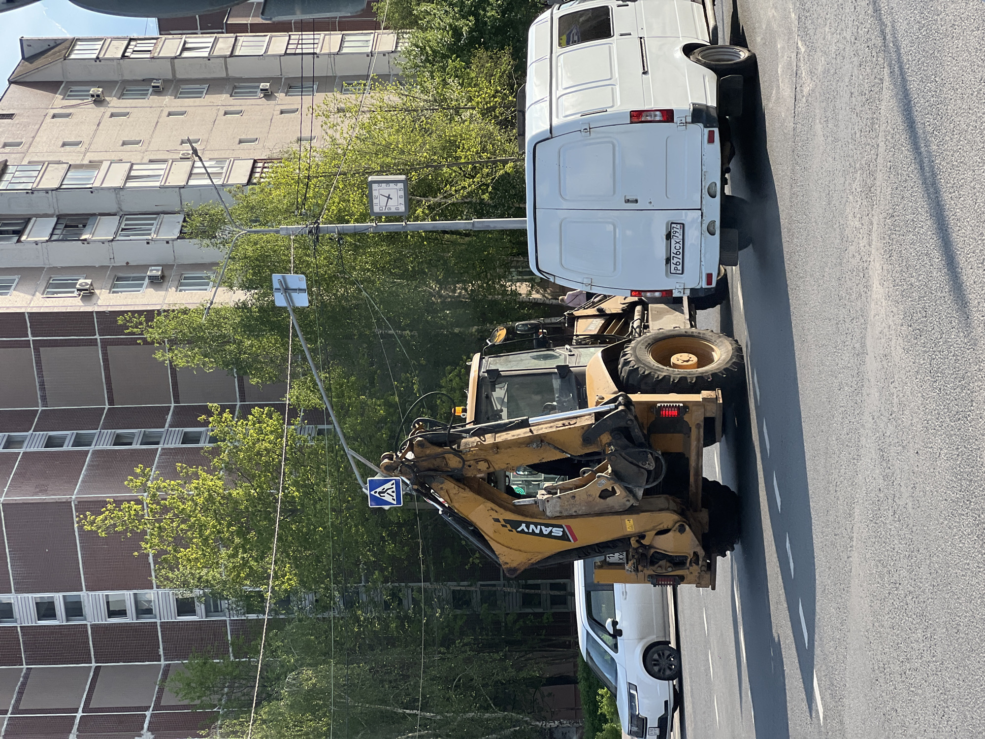 The image shows a street scene in Moscow, Russia. A yellow SANY backhoe is parked on the asphalt road, near a white van with the license plate P676CX797. A pedestrian crossing sign is near the backhoe. Above the vehicles are trees, power lines, and a clock on a pole. A large building with a patterned facade is visible in the background.