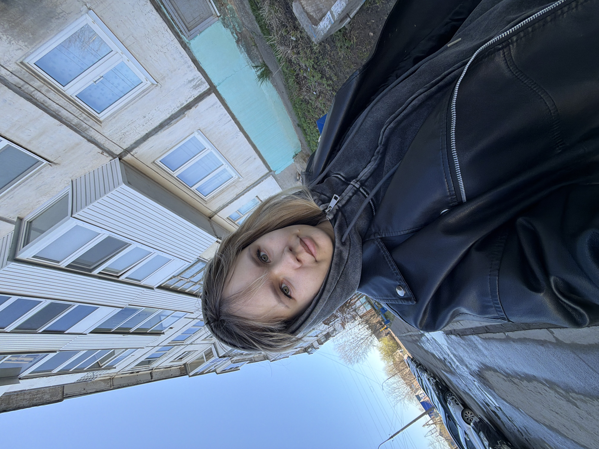 A young woman is standing outdoors in an urban environment. She is wearing a dark gray hooded sweatshirt and a black leather jacket. She appears to be facing the camera directly, with a slight tilt of her head.
Behind her, a multi-story building is visible. The building appears to be residential with a repetitive pattern of windows on each floor. Adjacent to the building is a strip of grass or vegetation. On the other side, there is an asphalt road with parked vehicles, suggesting a street scene. The sky is visible above the building.