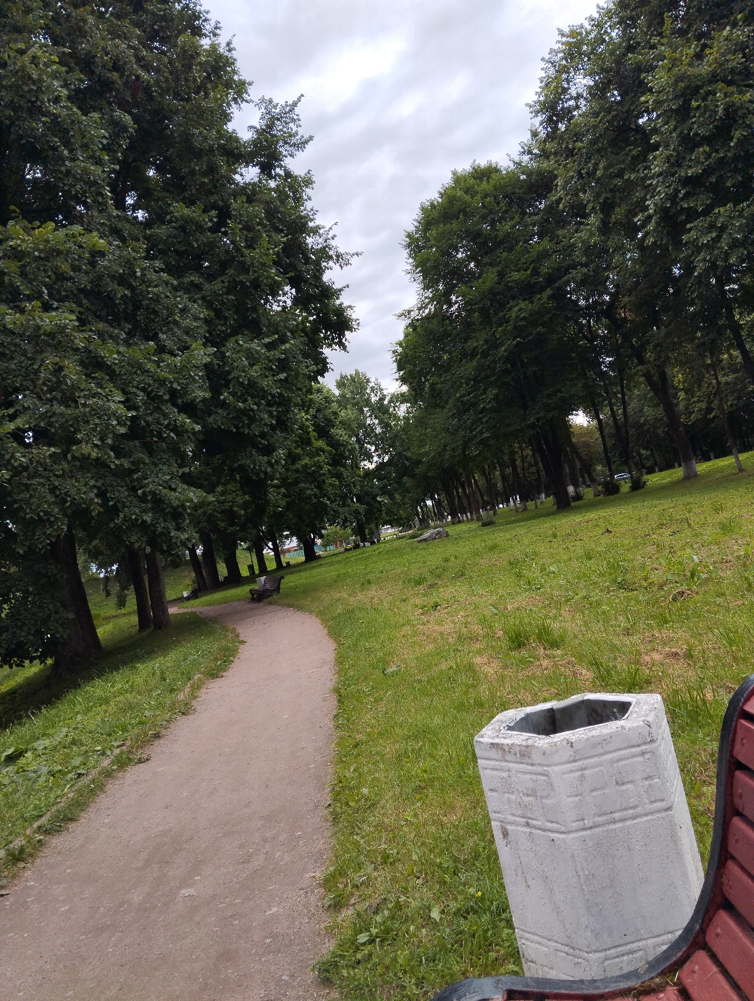The image depicts a park pathway in Kingisepp, Russia. A dirt path curves through a grassy area lined with mature trees. In the foreground, a concrete trash receptacle is visible next to a portion of a red park bench. The sky is overcast.