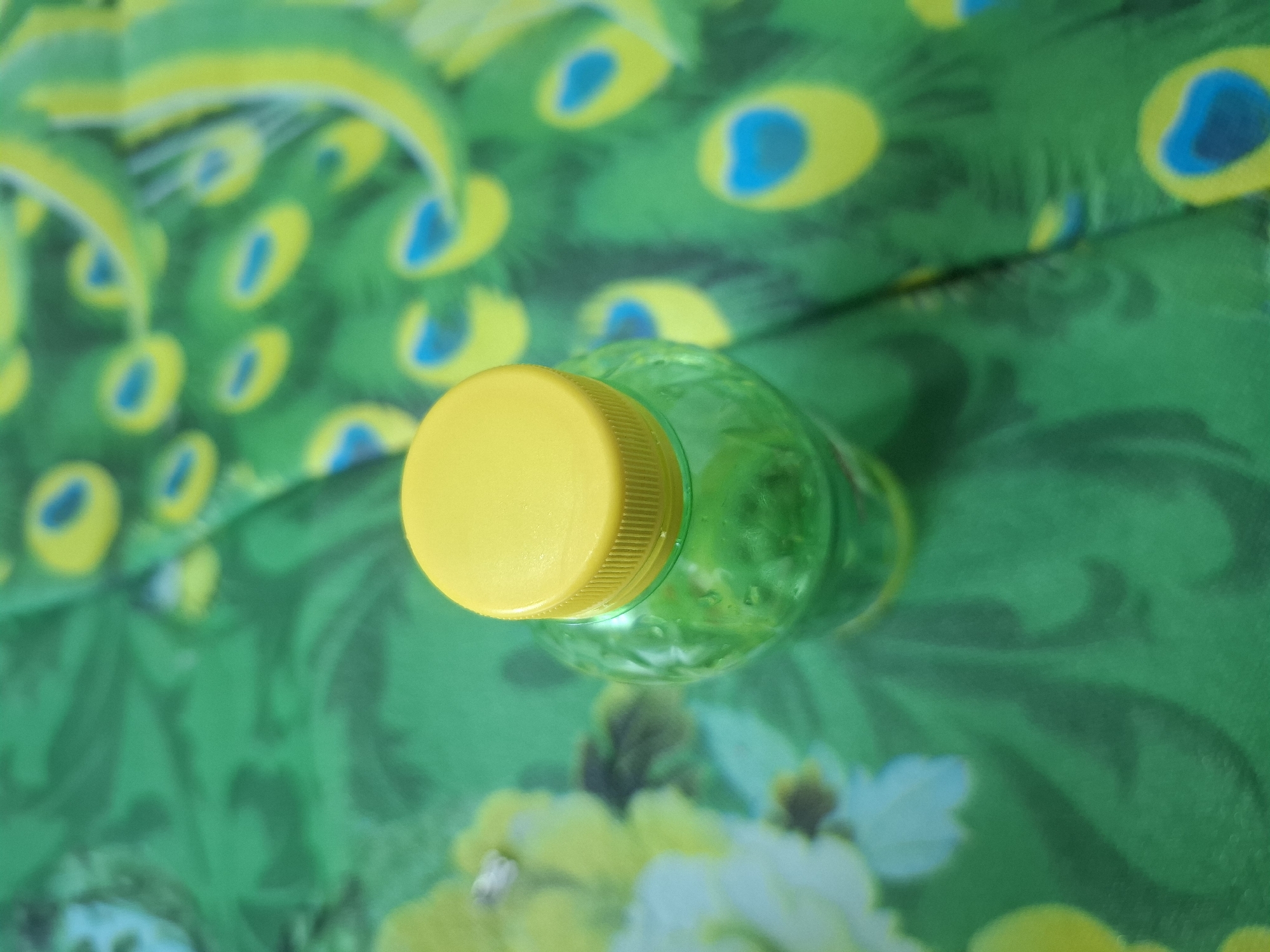 A green plastic bottle with a yellow cap sits on a green fabric with a peacock feather pattern. The bottle is translucent and appears to contain a green liquid. The fabric has a repeating pattern of peacock eye spots, with blue centers surrounded by yellow and green rings. The lighting is bright, casting a slight shadow beneath the bottle. There are no people or other objects visible in the image. This image was taken in Meureudu, Indonesia.