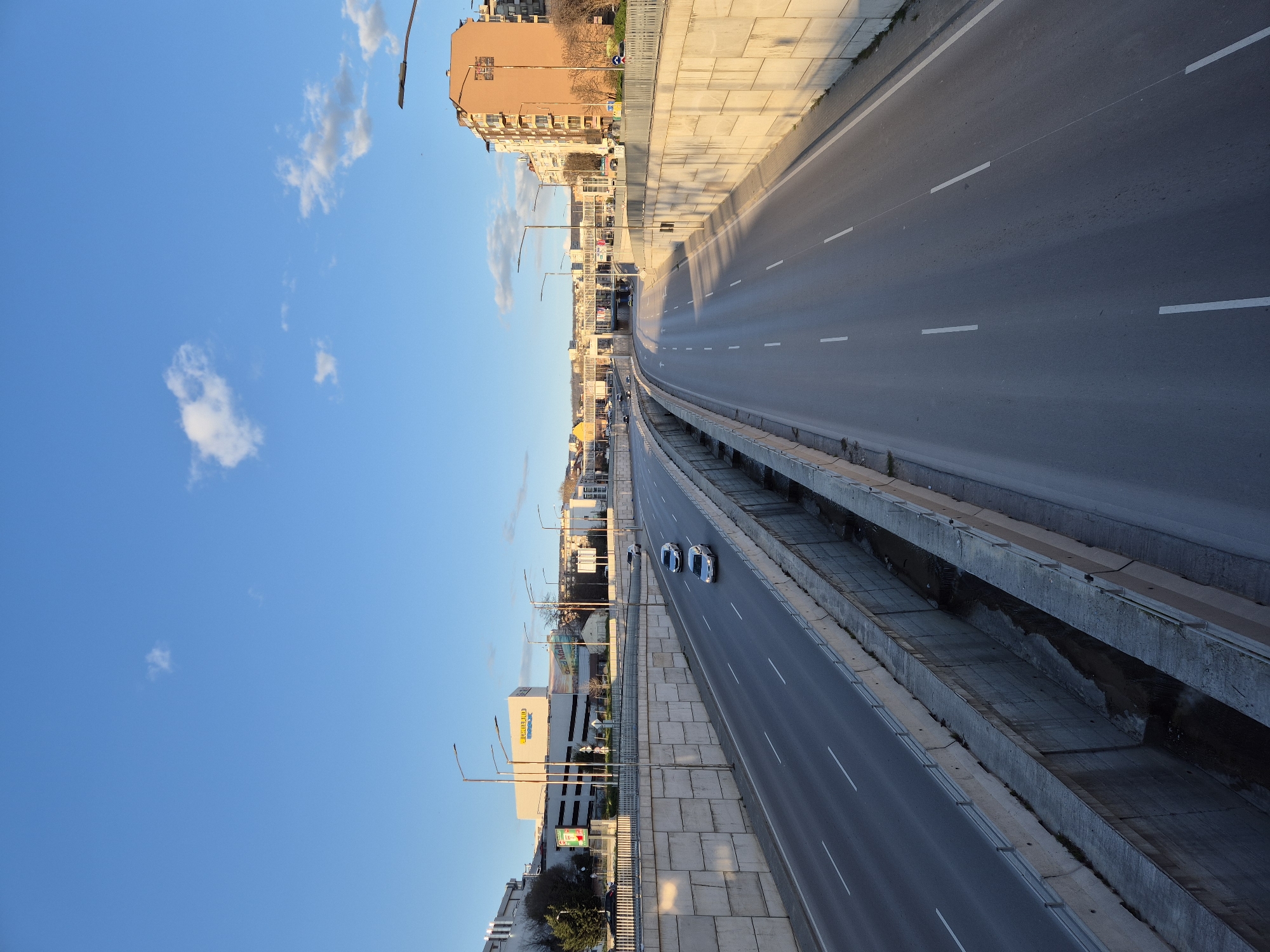 The image shows a highway running through the city of Varna, Bulgaria. Three cars are traveling on the highway. Buildings line one side of the highway, while a concrete barrier separates the highway from the background on the other side. The sky is blue with some clouds. Street lights line the side of the highway.