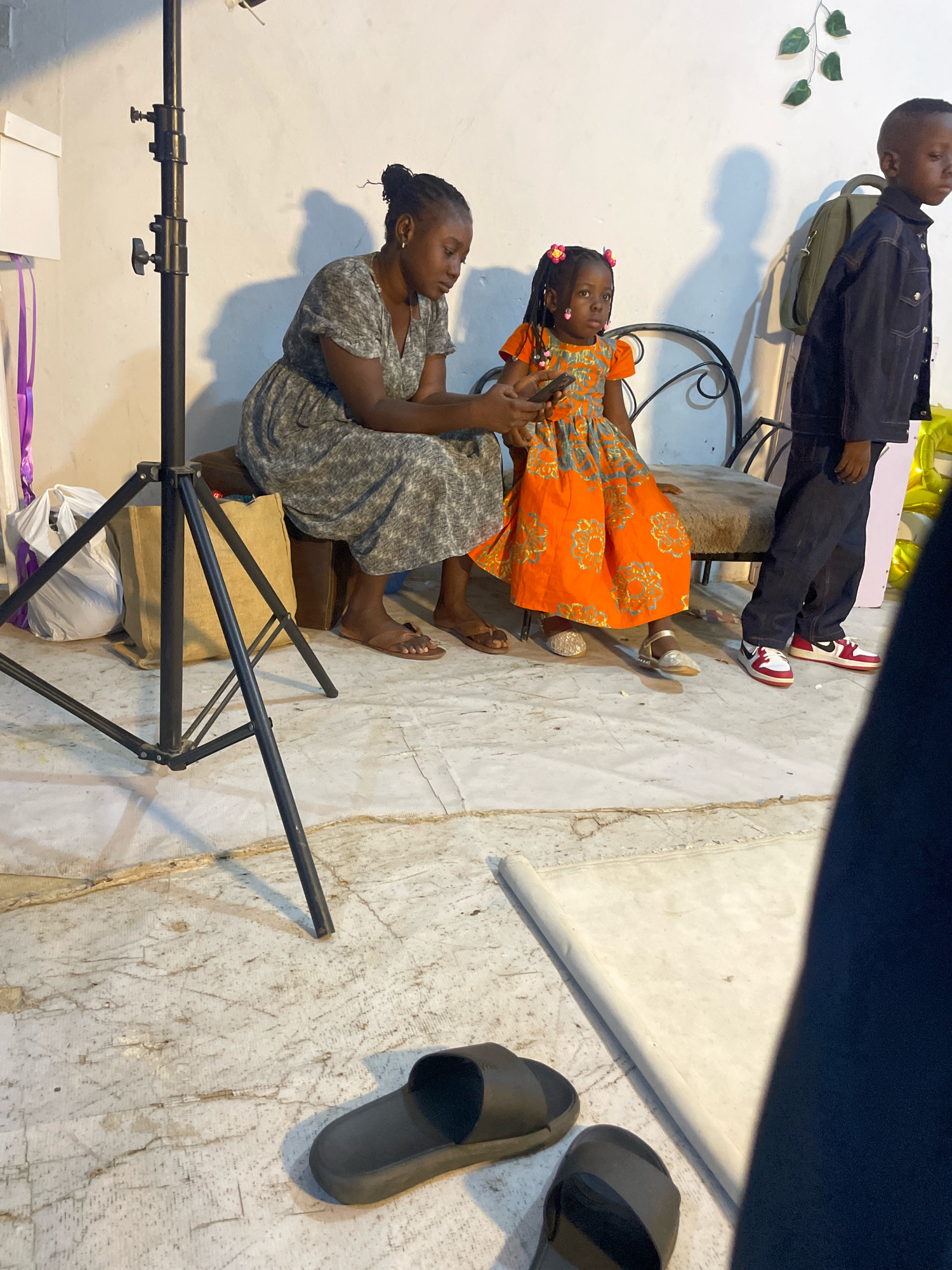 In an indoor setting, likely a residence or studio in Jos, Nigeria, three children and one adult woman are visible. The woman, wearing a grey printed dress, is seated on a dark brown cushioned bench, looking down at a mobile phone. Beside her, a young girl in an orange and grey patterned dress with pink hair clips is also seated, looking towards the viewer. To the right, a young boy in a dark denim jacket and jeans stands, facing away from the camera. In the foreground, a black light stand is positioned to the left, with a white bag and a burlap sack behind it. The floor is a light-colored, cracked concrete surface, with a pair of black slides and another dark shoe visible. A rolled-up white mat lies on the floor in the lower right quadrant, partially obscured by a dark blue garment. The wall behind the subjects is white, with a light green vine decoration and shadows cast on it.
