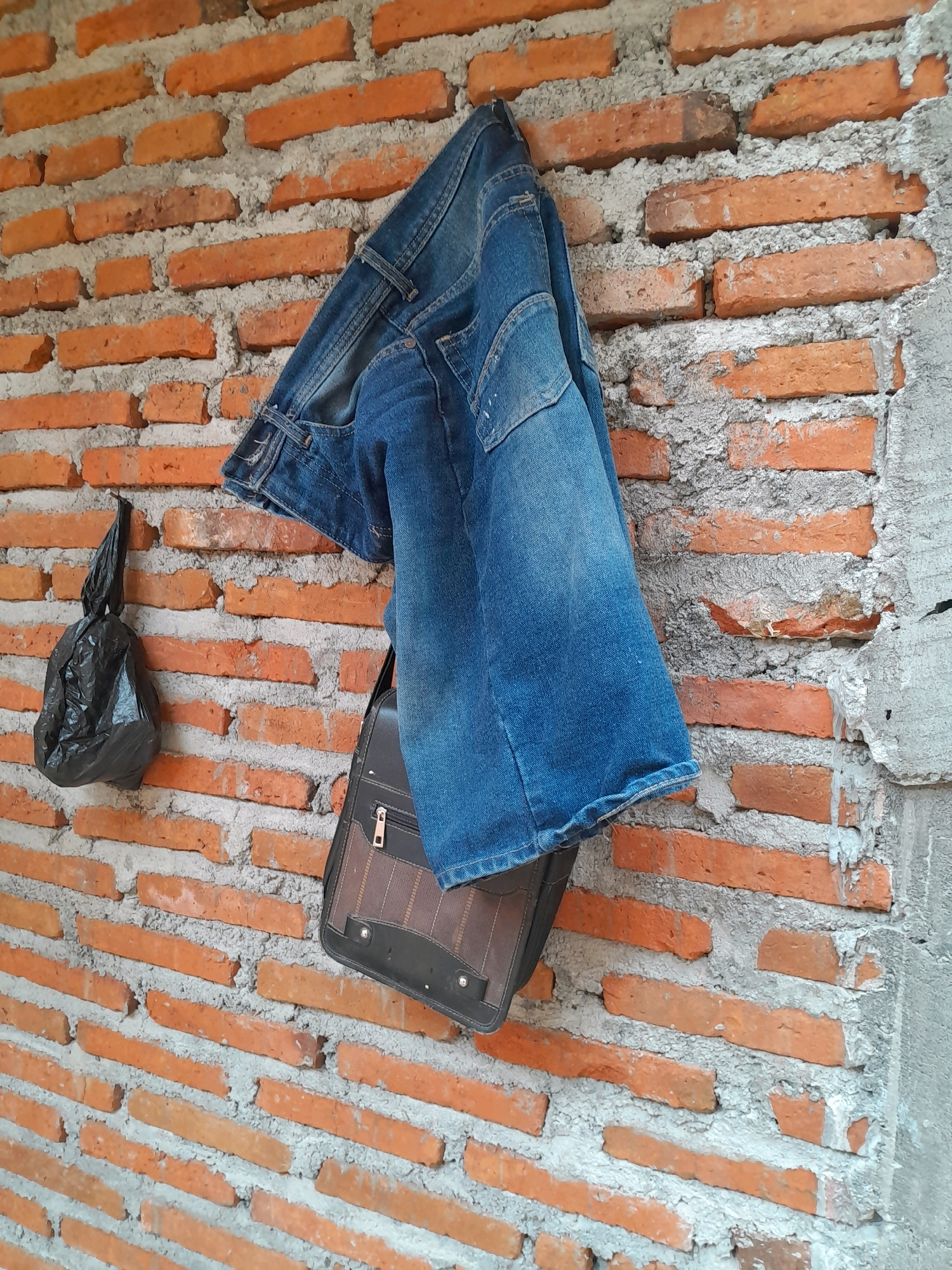 A close-up, eye-level shot shows a section of an exterior brick wall in Karangduren, Indonesia. The wall is constructed from rough, orange-red bricks with visible gray mortar. A pair of blue denim jeans is draped over a nail or hook, hanging from the wall. Below the jeans, a dark brown leather messenger bag with a zippered compartment and decorative stitching is also hanging. To the left of the jeans, a small, crumpled black plastic bag is suspended from a nail. The scene appears to be outdoors, possibly in a backyard or an alleyway, given the rough construction of the wall and the presence of discarded materials. The lighting suggests it is daytime.