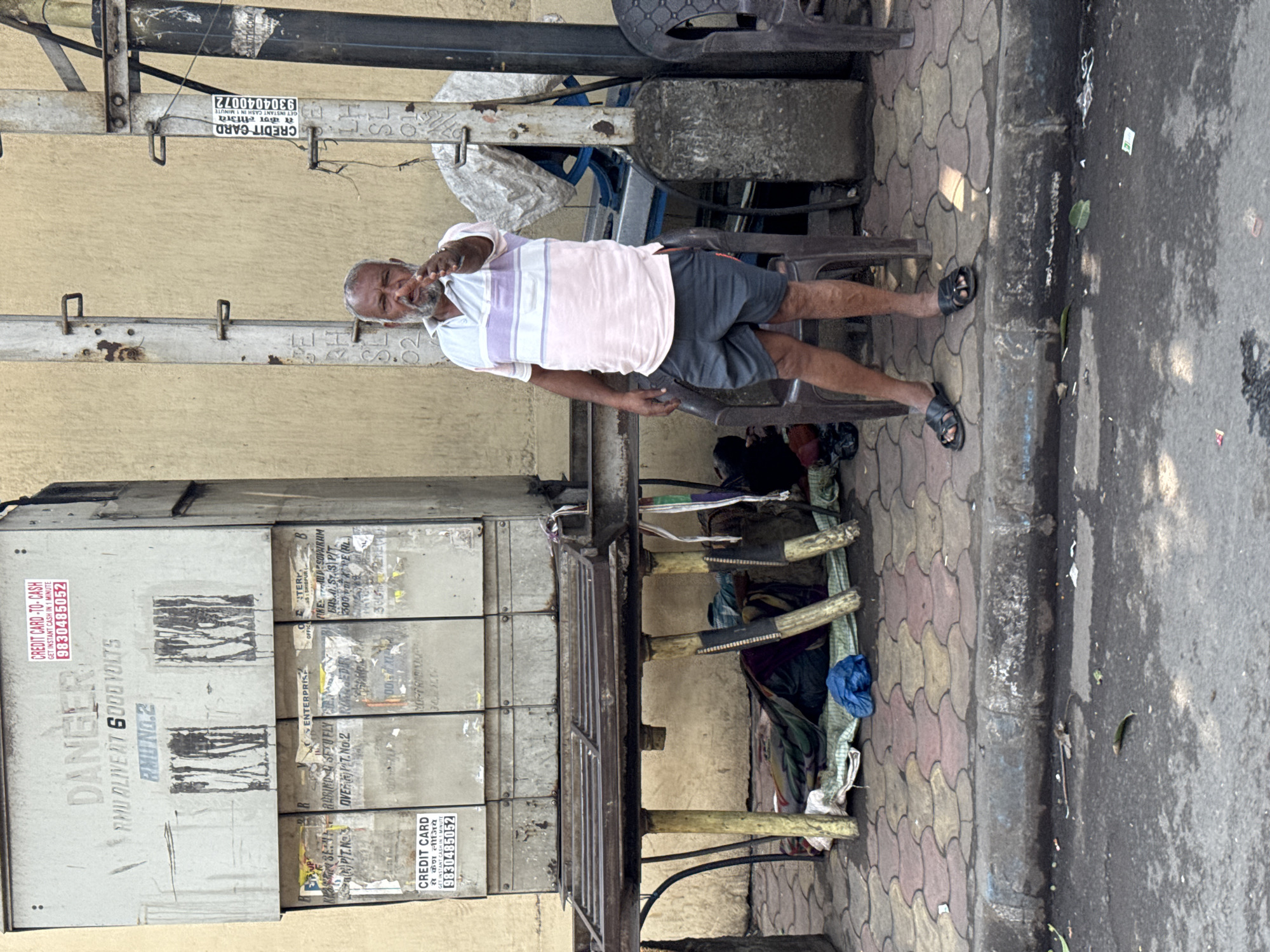 An elderly man stands on a paved sidewalk in an urban area of Kolkata, India, pointing directly at the camera. He has a short grey beard and hair, and wears a pink, white, and light blue striped polo shirt, dark shorts, and black sandals. He is positioned slightly right of center, in front of a dark plastic chair.
To his left, a large, weathered grey electrical distribution box or transformer is affixed to a wall. It bears the stenciled warnings "DANGER" and "RMU ALIVE AT 6000 VOLTS," along with "RMU NO.2." Multiple torn advertisements are visible, including "CREDIT CARD - TO - CASH GET INSTANT CASH IN 1 MINUTE" with phone number 9830485052. Beneath this box, a makeshift sleeping area is arranged on the ground under a metal table-like structure. It consists of various folded fabrics (green, maroon, dark blue) serving as bedding, with a blue plastic bag nearby.
To the man's right, two tall, corroded grey metal poles stand against a light beige wall. The rightmost pole also displays a "CREDIT CARD - TO - CASH" advertisement with phone number 9804040072. Behind this pole, a blue object is partially covered by a white tarp, and another dark plastic chair is visible to the far right.
The sidewalk is paved with interlocking tiles in shades of red, yellow, and grey. A concrete curb separates the sidewalk from a dark asphalt street in the foreground, which shows scattered debris including leaves and small pieces of litter. The scene is brightly lit, indicative of daytime.