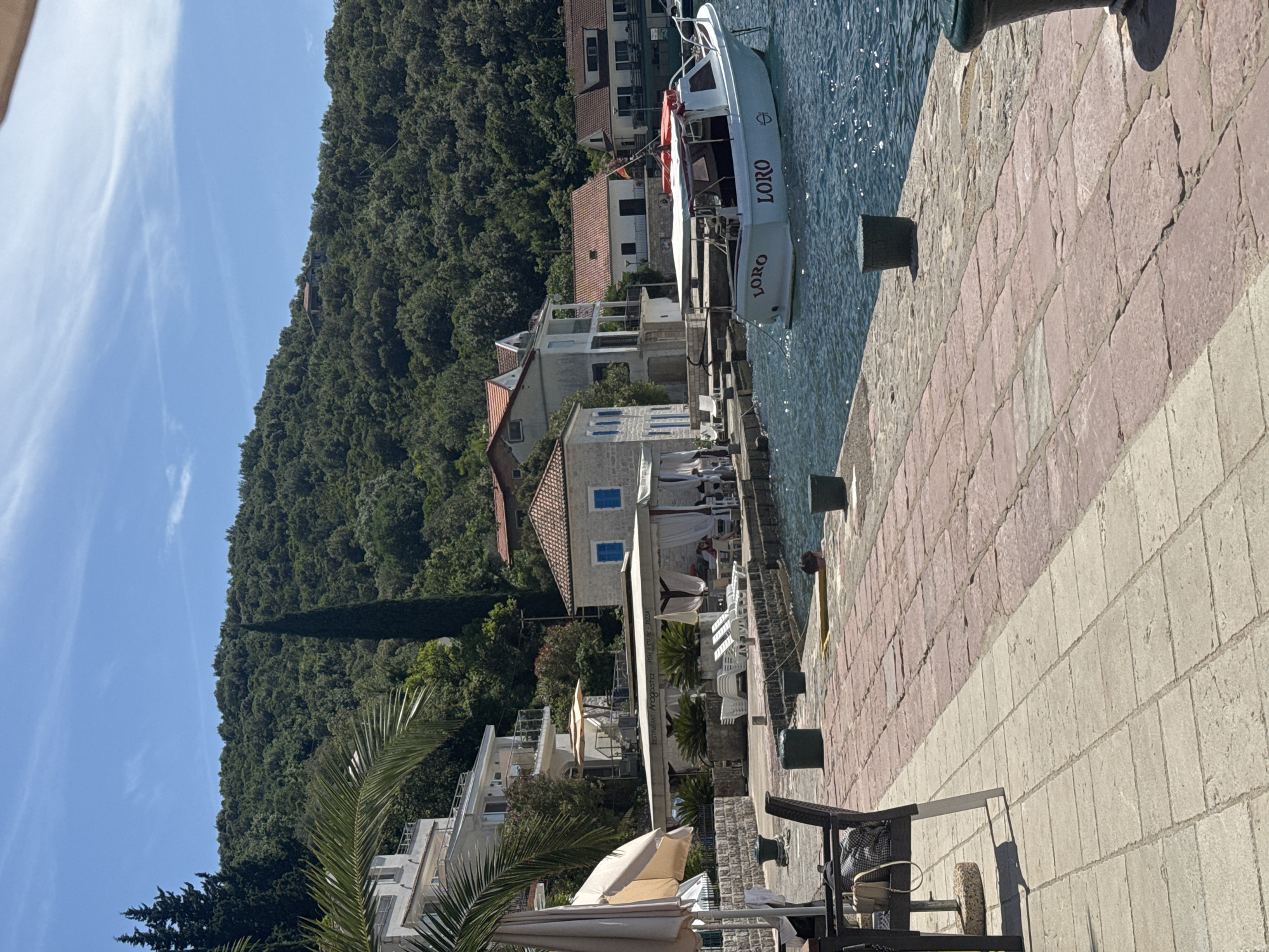 The image depicts a sunlit waterside scene in an unspecified coastal settlement. In the foreground, a two-toned paved promenade consists of light-grey square pavers transitioning to reddish-brown irregular pavers. A dark outdoor chair with a checkered cushion and a light-colored handbag rests on the left. Several dark bollards are spaced along the promenade's edge.
The midground features calm, blue-green water where a white motorboat, named "LORO" in red capital letters on its stern and starboard side, is moored. A person's head is visible in the water near a yellow float behind one of the bollards. Adjacent to the promenade, a restaurant named "Restaurant Aragogsta" with white canopies offers outdoor seating, including white lounge chairs. Behind it are several buildings: one white building with blue window shutters and a red-tiled roof, and other multi-story white buildings with balconies and glass railings. Palm fronds extend into the left side of the frame.
The background is dominated by steep, densely forested hills covered in various shades of green trees, including a prominent tall, slender cypress-like tree. Scattered residential structures with red-tiled roofs are visible amidst the foliage on the hillsides. The sky above is clear and bright blue with faint, wispy white clouds. The overall lighting suggests a sunny day.