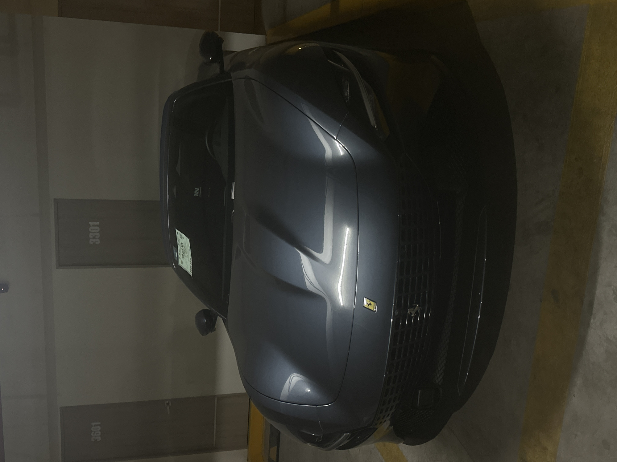 The image shows the front of a dark gray Ferrari parked in an indoor parking garage. The car is angled, taking up most of the frame. The Ferrari logo is visible on the grille. Behind the car, apartment or storage unit doors are visible, labeled "3301" and "3601." Yellow parking lines are painted on the floor to the car's right. A temporary sticker is visible on the windshield. There is no evidence to suggest a specific location in Puebla, Mexico. There are no people visible.