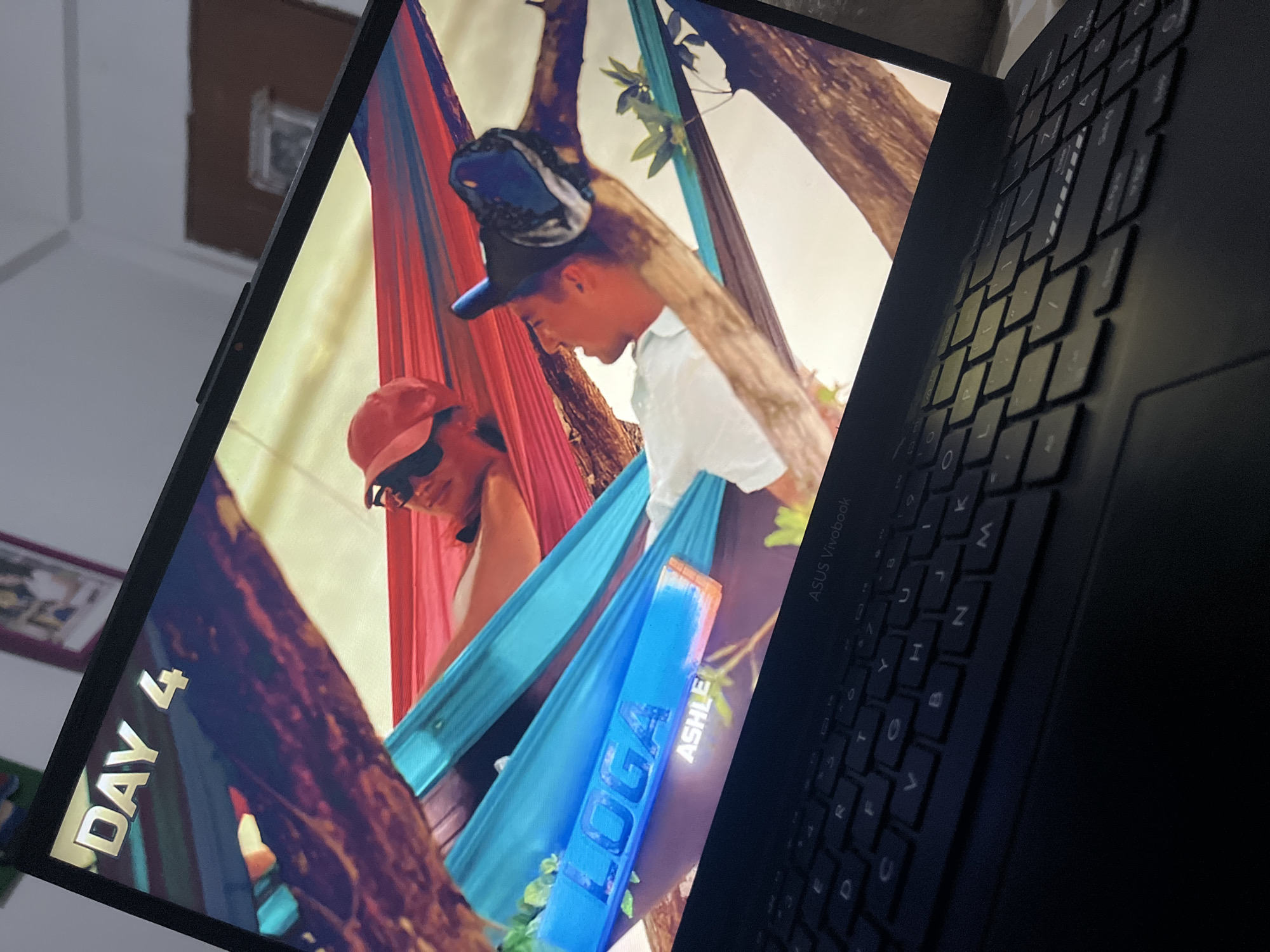 An ASUS Vivobook laptop is depicted from a low, angled perspective, with its screen displaying a still frame from a video. The screen features two men lying in hammocks outdoors, suspended between tree branches. The man on the left wears a red baseball cap and appears to be wearing sunglasses while reclining in a red hammock. The man on the right, wearing a blue and black baseball cap, is in a turquoise-blue hammock. Both men are positioned facing towards each other. In the upper-left corner of the screen, the text "DAY 4" is displayed in a metallic, three-dimensional font. In the lower-left area, a blue graphic overlay contains the name "LOGA" in prominent white letters, with "ASHLE" (presumably Ashley) visible beneath it in a smaller white font. The background of the video footage shows natural outdoor elements, including trees and a bright sky. Reflections on the laptop screen reveal elements of the surrounding indoor environment, including a light-colored wall, faint outlines of framed items, and a ceiling. The laptop's black keyboard and trackpad are visible in the foreground, and the "ASUS Vivobook" brand name is present above the keyboard on the right side. The image contains no discernible visual information or landmarks to indicate a specific location in Santa Ana, Venezuela.