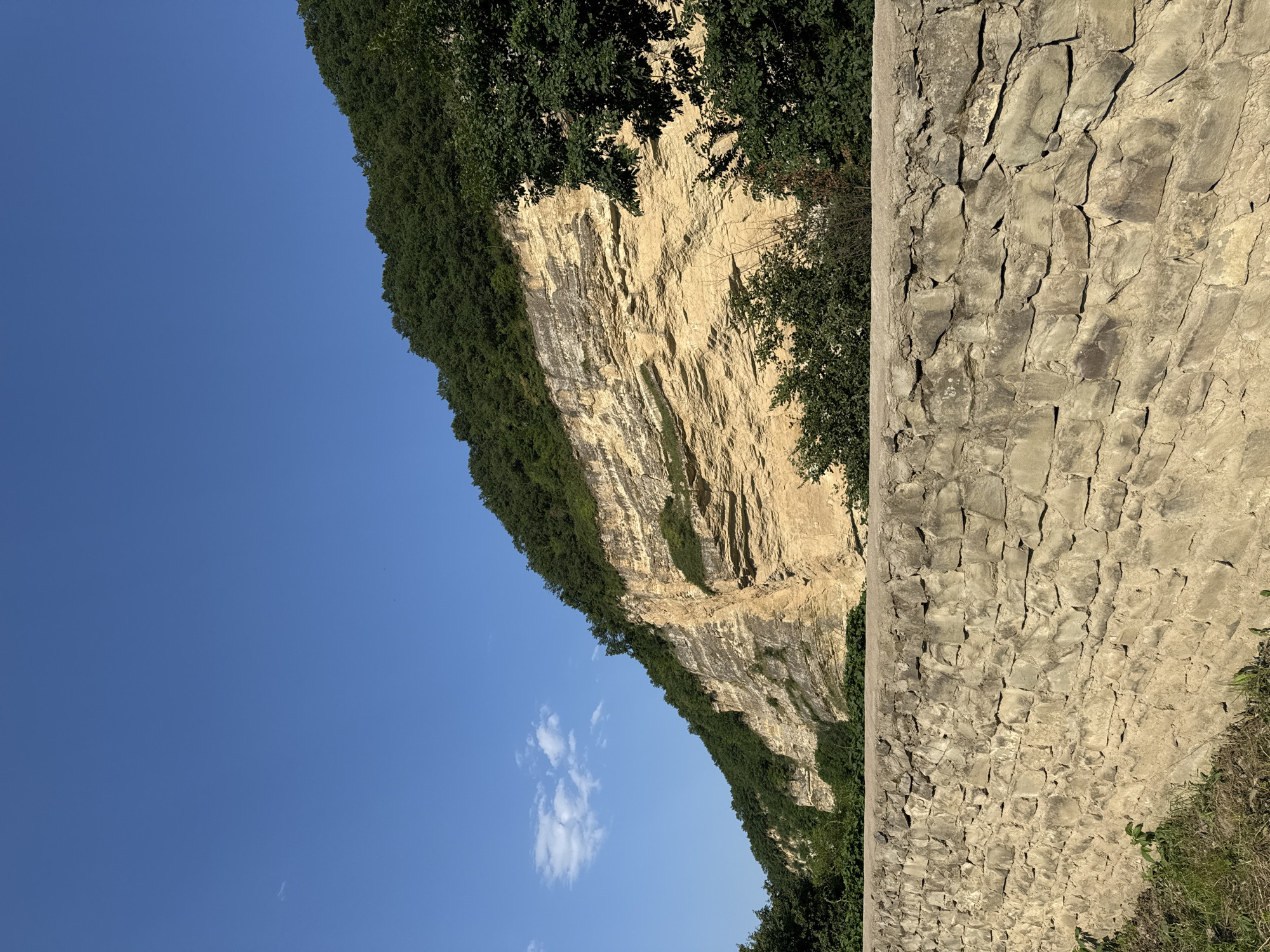 A high-angle, medium shot captures a natural landscape beneath a clear blue sky with a few wispy clouds. In the foreground, a rustic stone wall runs horizontally across the frame, constructed from irregular, light-colored stones with visible mortar. Green grass and weeds grow along the base of the wall. Beyond the wall, a steep, layered rock face dominates the middle ground, its warm, sandy-yellow tones contrasting with the dark green foliage that covers its upper slopes and the surrounding trees. The rock face exhibits distinct horizontal striations, indicating geological layers, with patches of greenery growing from crevices. The scene is located in the village of Khapil, Russia, suggesting a natural, mountainous or hilly terrain.