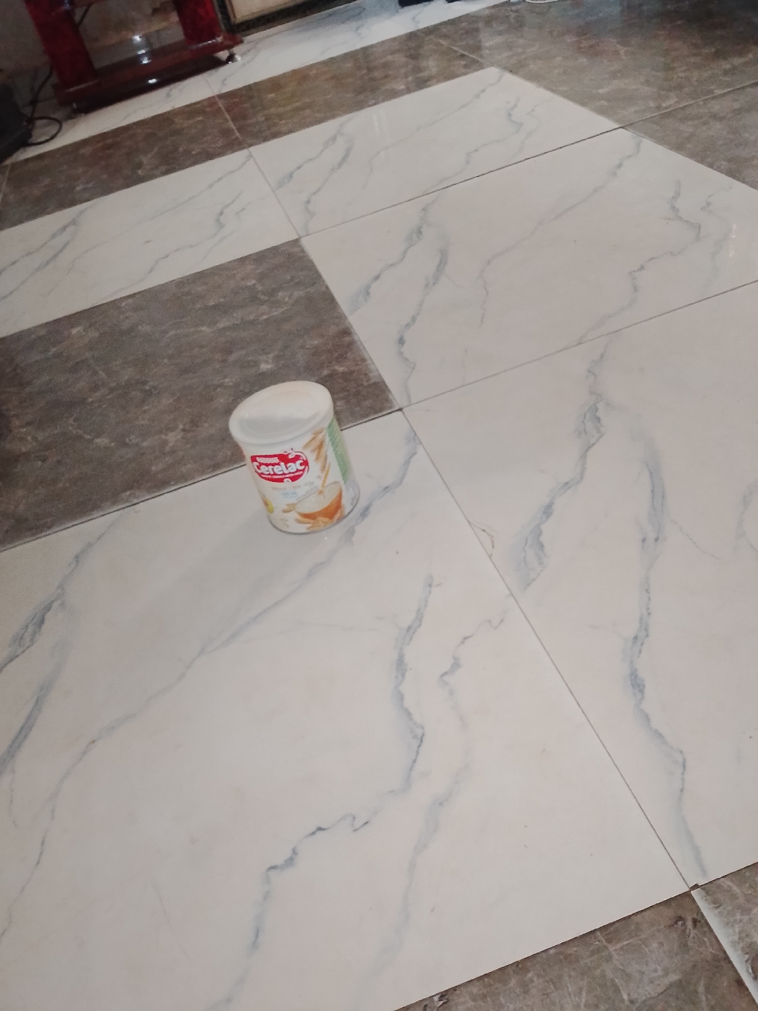 A white, cylindrical can of Cerelac infant cereal is placed on a floor covered with large, rectangular marble-patterned tiles. The tiles are a mix of white with grey veining and a darker grey solid color, arranged in a checkerboard pattern. The can is positioned on a white tile with grey veins, slightly tilted. The scene appears to be indoors, likely in a residential setting in Jalingo, Nigeria. The lighting suggests an ambient interior light source. No people are visible in the image.