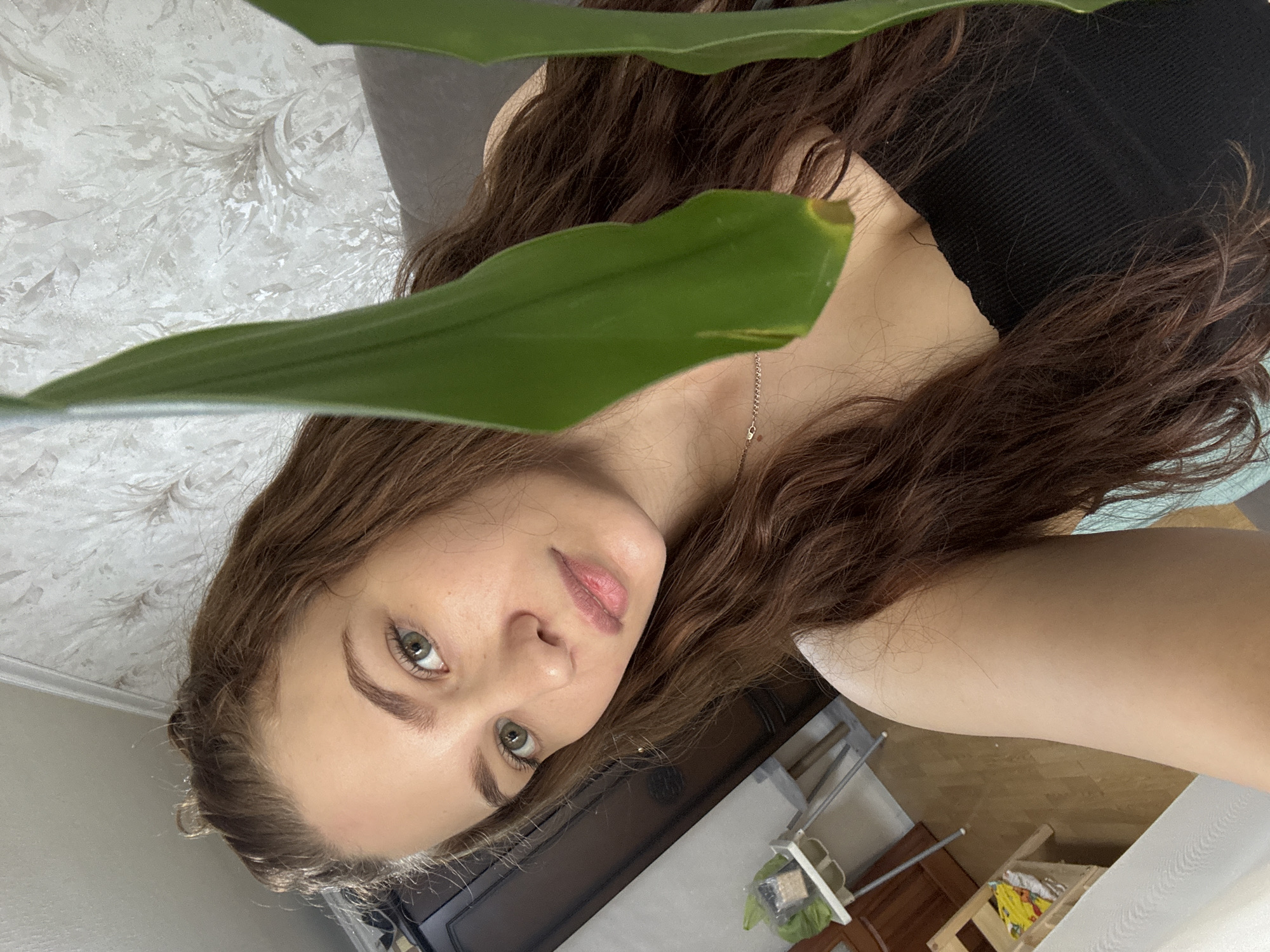 The media file is a close-up indoor shot of a young woman, rotated 90 degrees counter-clockwise.
The primary subject is a young woman, whose face and upper body are partially visible, framed and somewhat obscured by large, vibrant green leaves of a houseplant in the immediate foreground. She has long, wavy brown hair, light-colored eyes (appearing greenish-hazel), and fair skin. She wears a dark top and a delicate, thin necklace. Her gaze is directed straight at the camera with a neutral, slightly contemplative expression.
The scene depicts an indoor setting, likely a home. In the background, to the left of the woman's head, there is a wall covered with light-colored wallpaper featuring a subtle, textured pattern of grey or silver floral/swirl motifs. To the right, various pieces of furniture are visible, including a dark wooden cabinet or shelf, and a lighter wooden structure resembling a high chair or stool. Indistinct items are placed on and around this furniture. The flooring in this area appears to be light wood or laminate.
The activity captured is a self-portrait or a posed photograph, with the woman looking directly at the lens. The lighting suggests it is daytime. No text is visible in the image. The location context is Chaikovsky, Russia.