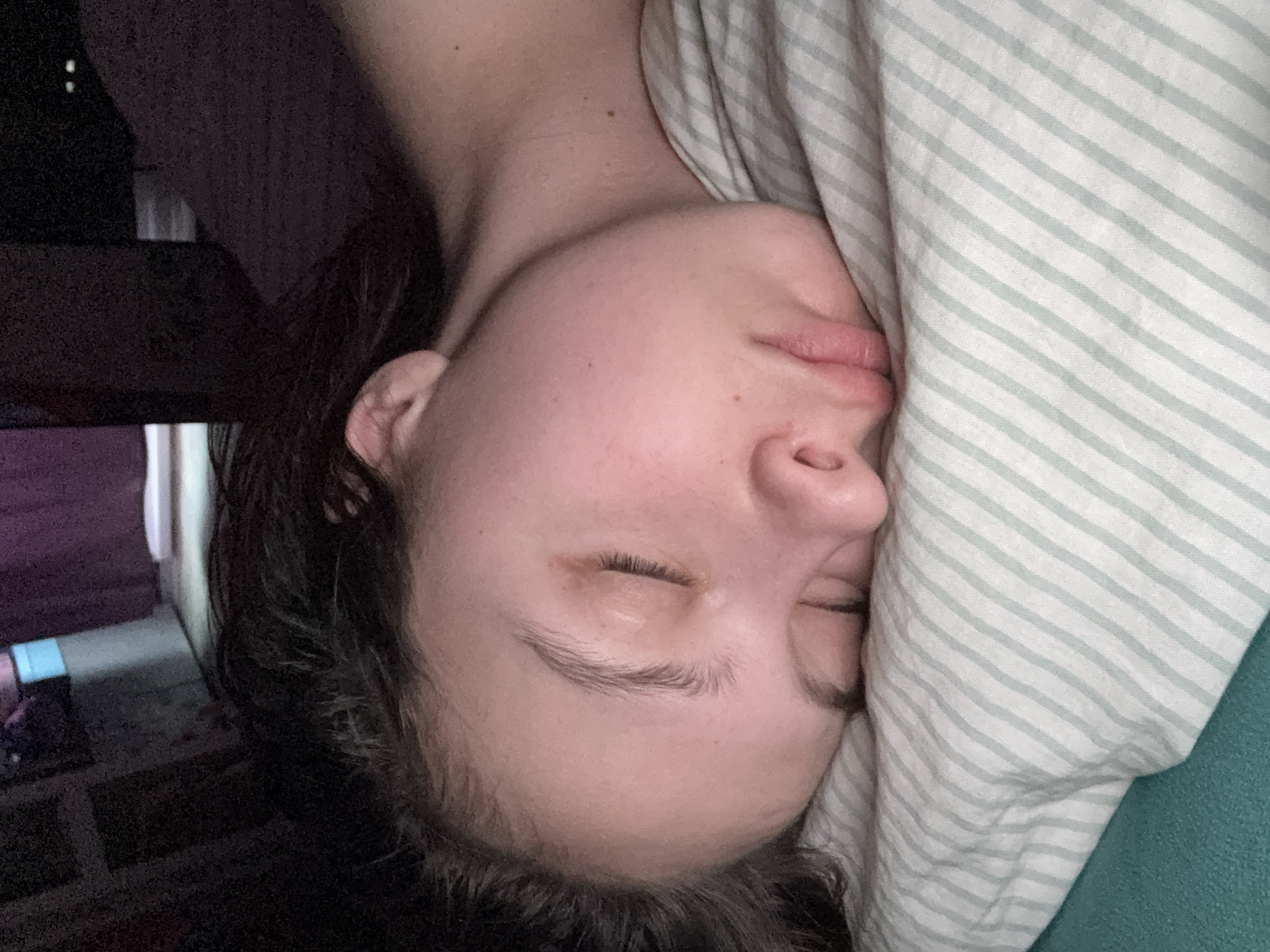 A close-up, tilted shot depicts a person sleeping. Their face, skin tone light, is pressed against a striped pillow. The person's eyes are closed and their dark hair is visible. In the background is some furniture. The location is Прикамский, Russia.