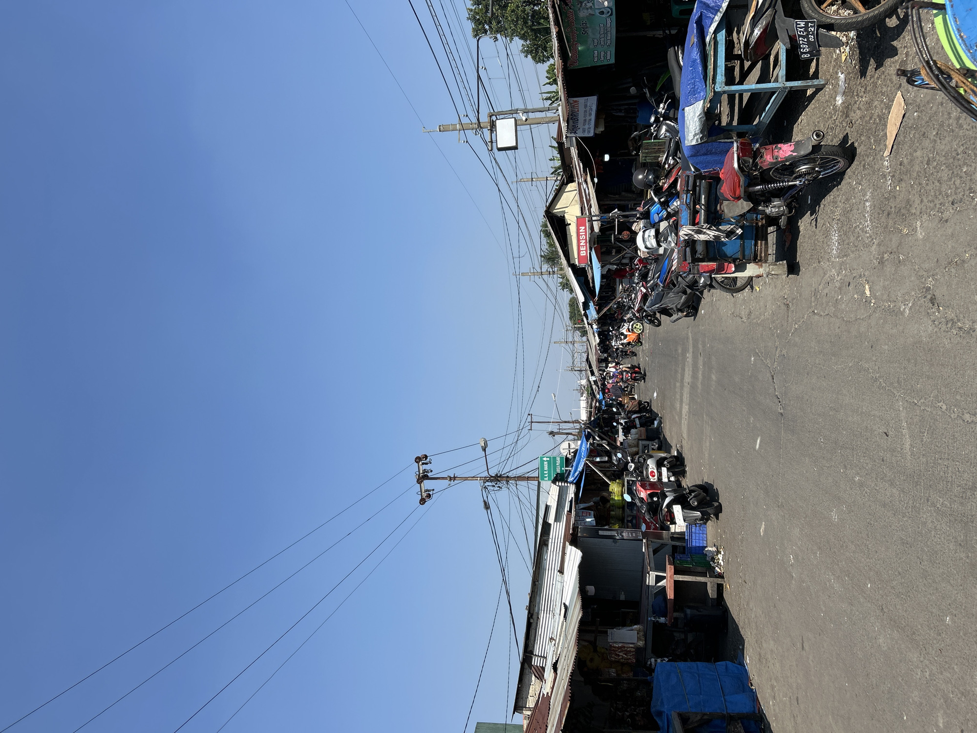 This is an eye-level, outdoor, medium shot of a bustling street market in Depok, Indonesia. The street is paved and lined with open-air stalls and shops, many with corrugated metal roofs. A clear blue sky is visible above. A prominent feature is the abundance of parked motorcycles and scooters on both sides of the street, suggesting it is a common mode of transportation and that the market attracts many visitors. There are a few people visible, mostly walking or tending to their stalls, but their faces are not clearly discernible. Some of the stalls appear to be selling goods such as food items, household supplies, and possibly clothing. Signs in Indonesian are visible on some of the stalls, including one that says "BENSIN," indicating fuel is sold there. Power lines crisscross the sky, supported by utility poles, which are characteristic of urban infrastructure. The overall impression is a lively and active marketplace environment, typical of a local market in Indonesia.