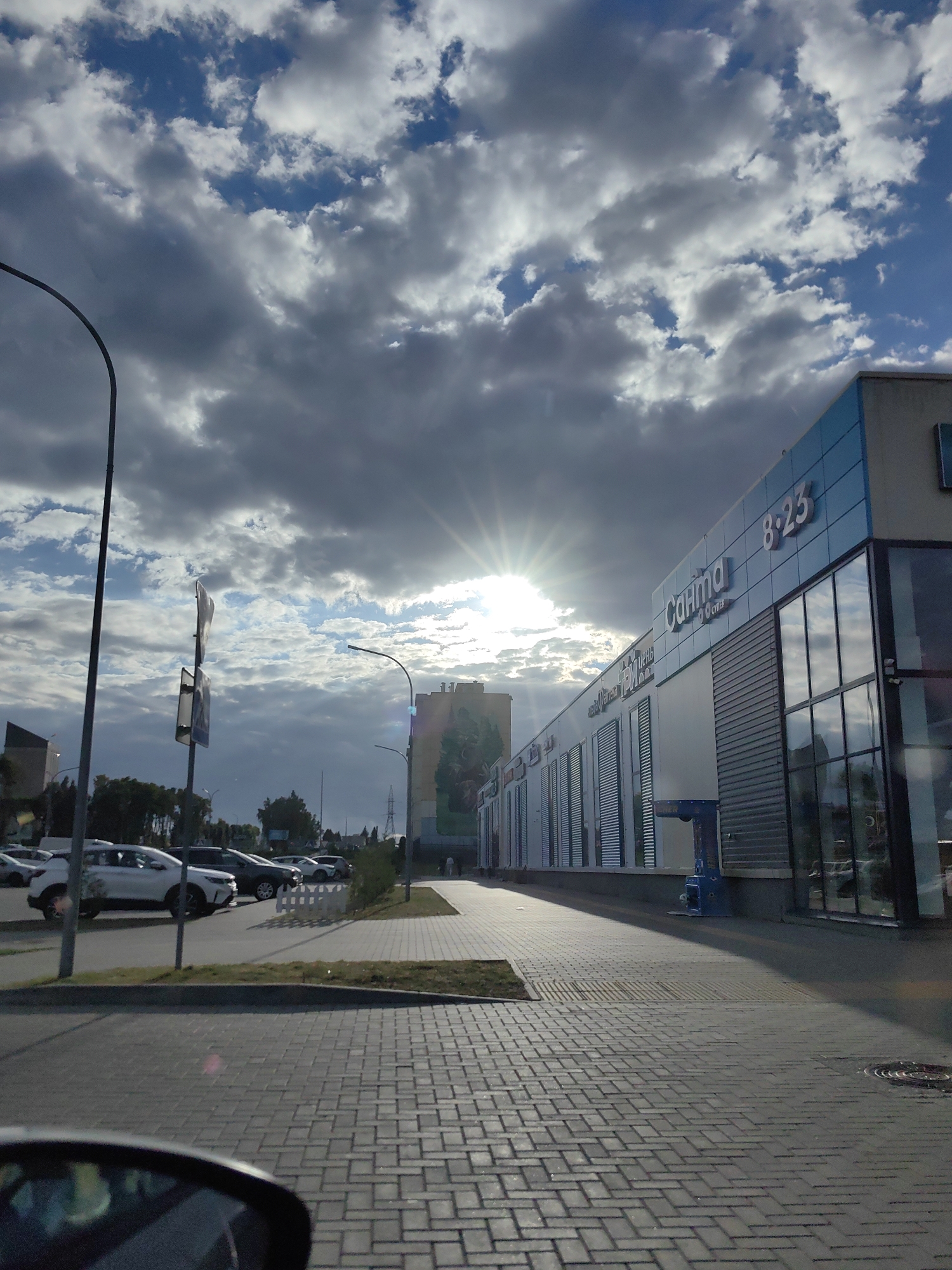 The image depicts a street scene in the city of Dzyarzhynsk, Belarus, on a partly cloudy day. The sun is partially obscured by clouds, creating a sunburst effect. A modern, light blue building with large glass windows and horizontal siding is prominently featured on the right side of the frame. Signage on the building includes the word "Санита" and the numbers "8:23". In front of the building, a paved walkway is visible, leading away from the viewer. To the left, a parking lot with several cars is seen. A street lamp stands on the left side of the image, and further back, other buildings and trees are visible. The ground in the foreground is paved with rectangular bricks laid in a herringbone pattern. A car's side mirror is partially visible in the bottom left corner of the frame.