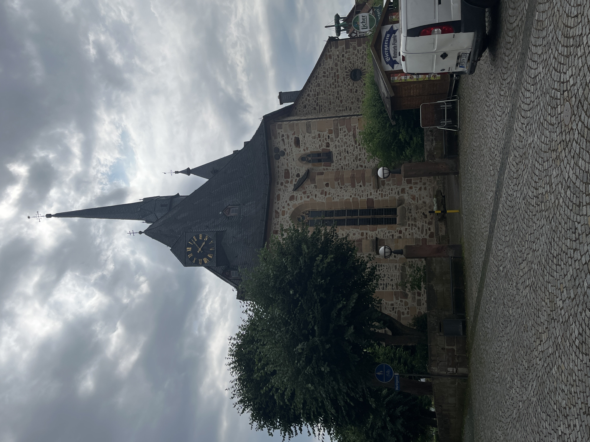 This image depicts a view of a stone church in Neustadt, Germany, under an overcast sky. The church has a prominent clock tower with Roman numerals and dark, steep roofs. A tall, pointed spire with a weather vane rises from one section of the roof. In the foreground, a large, leafy tree partially obscures the lower part of the church. To the right of the church, a white van is parked next to a small building with signs that read "Biergarten" and "Ristorante Pizzeria". The ground is paved with cobblestones. The lighting suggests it is daytime, likely late afternoon, given the cloudy conditions. The overall atmosphere is somewhat muted due to the weather. There is a street sign visible near the tree that reads "Marktgasse".