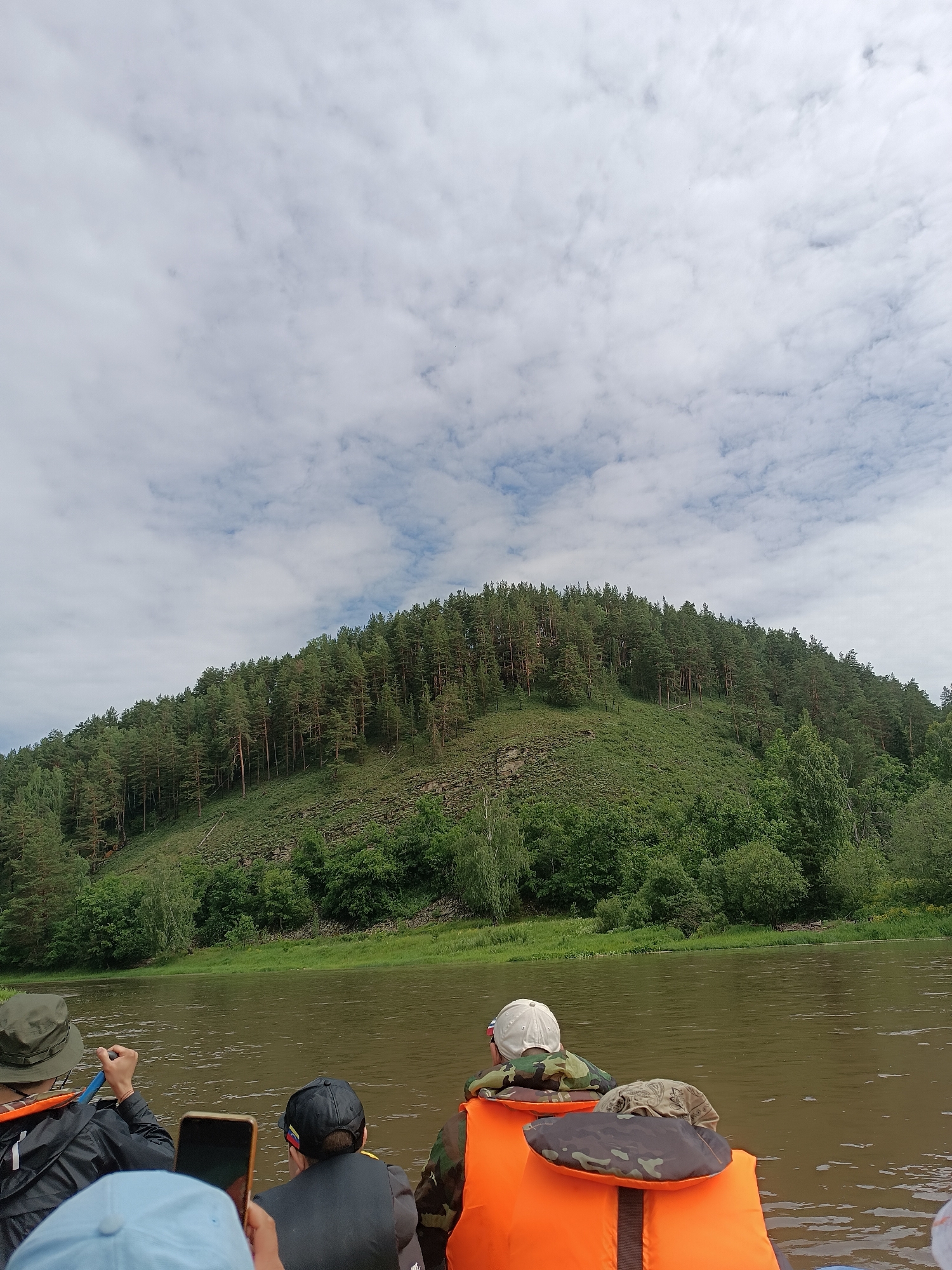 The image depicts a serene river scene with a densely forested hill rising from the opposite bank. In the foreground, three individuals are seated in a boat, facing away from the camera. The person on the left wears a dark jacket and a khaki hat, holding a paddle. The middle person is attired in a camouflage jacket and a black baseball cap, while the person on the right is also wearing a camouflage jacket and an orange life vest. The river water is a murky brown, reflecting the overcast sky. The forested hill is a mixture of deciduous trees and tall, slender pines. The location is identified as the city of Sergeevka, Russia.