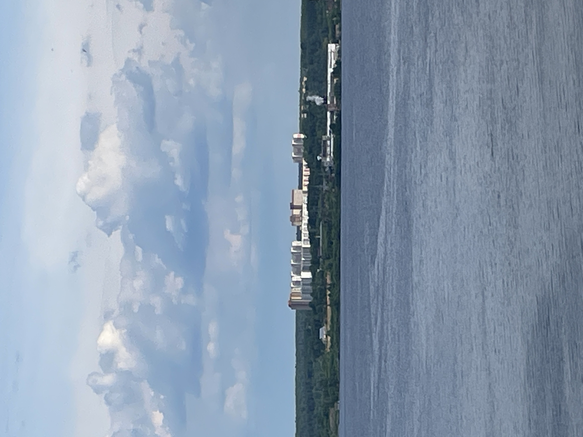 The image captures a wide, serene landscape view across a large body of water towards a distant shoreline under a partly cloudy sky. The scene is presented during daytime, with clear visibility and diffuse sunlight.
The right side of the frame is dominated by a vast expanse of calm, blue-gray water, showing subtle ripples across its surface that reflect the sky above.
Across the middle of the image, defining the horizon, is a distant landmass. This shoreline is densely covered with dark green trees, suggesting a forested or heavily vegetated area. Above the tree line, a row of multi-story buildings is visible, forming a small urban or suburban cluster. These structures are predominantly light-colored, suggesting modern residential or commercial architecture. Towards the right side of the building cluster, faint plumes of white smoke or steam rise from what appears to be an industrial facility or power plant situated on the distant bank.
The left side of the image features a sky filled with large, bright white cumulus clouds set against a lighter blue sky. No people or distinct activities are visible in the tranquil scene. The image provides a panoramic perspective of a quiet waterside environment, consistent with locations along major waterways, such as in "Пятый километр, Russia." No visible text is present in the media.