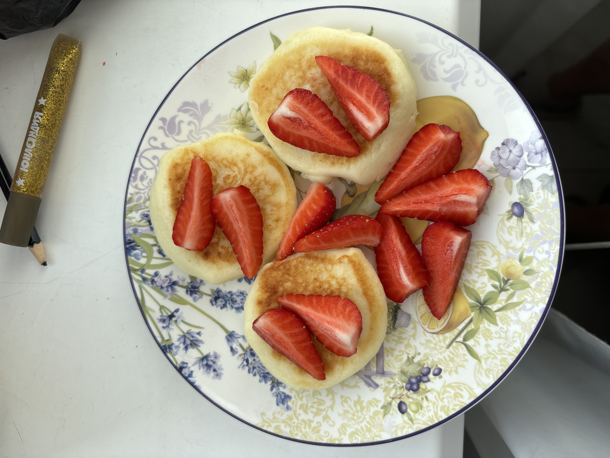 A top-down shot depicts a plate of three round, golden-brown pancakes topped with sliced strawberries, located in an unidentified city and state. The plate features a floral pattern with blue lavender and yellow lemons. A gold glittery pen is positioned to the left of the plate. The background is a plain white surface, possibly a table, with a hint of white and gray suggesting the presence of a chair or other furniture in the lower right corner. The scene is well-lit and emphasizes the pancakes and strawberries.