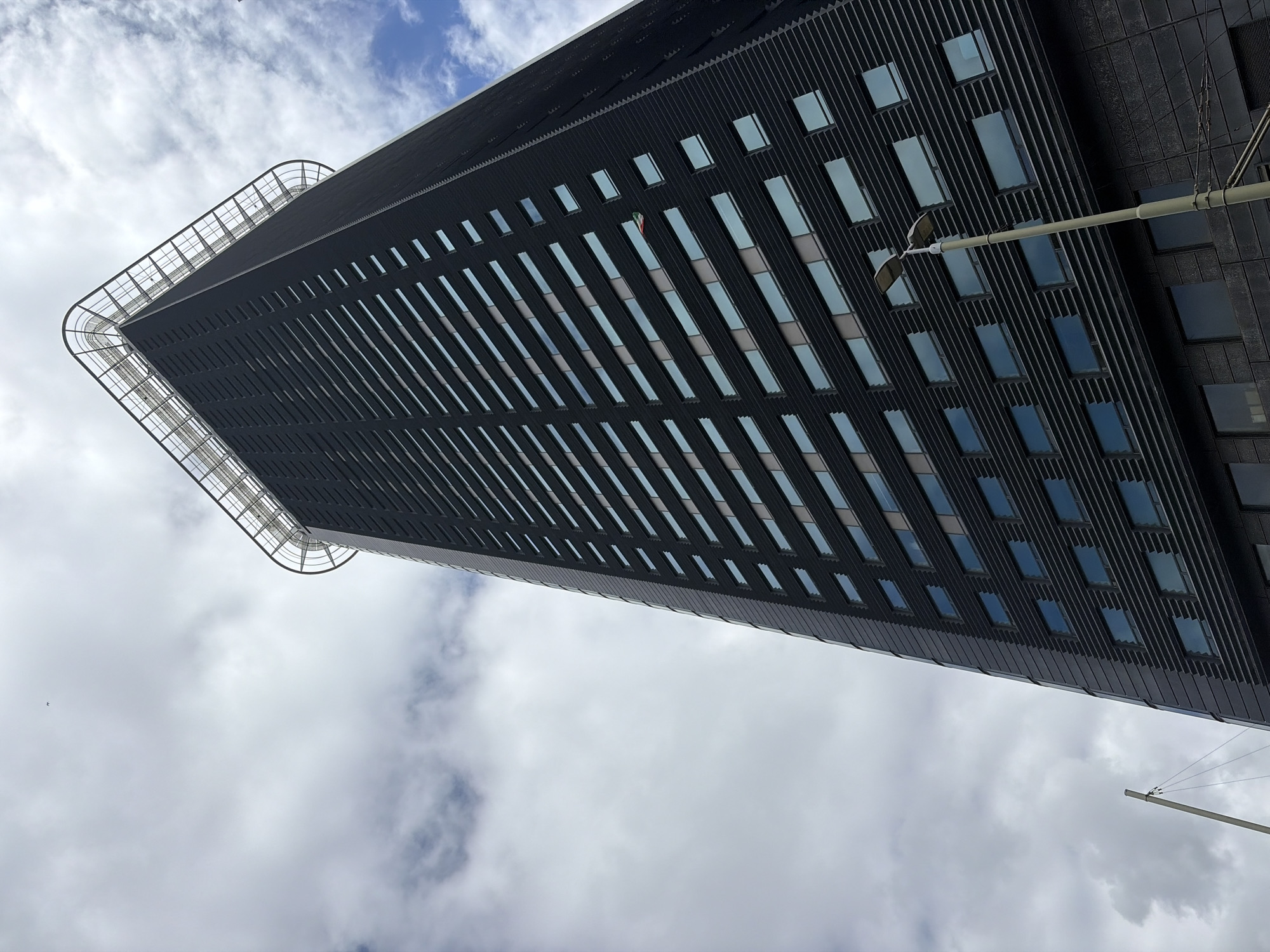 The photograph showcases the exterior of a tall, dark, modern skyscraper in The Hague, Netherlands. The building is viewed from a low angle, emphasizing its height and imposing structure against a cloudy sky with patches of blue. The skyscraper features a facade composed of dark, textured panels interspersed with numerous rectangular windows. At the very top of the building, a prominent helipad structure with a metal railing is visible. A streetlamp on a pole stands in the foreground, positioned to the right of the frame and extending upwards. The overall impression is one of urban architecture under a dynamic sky.