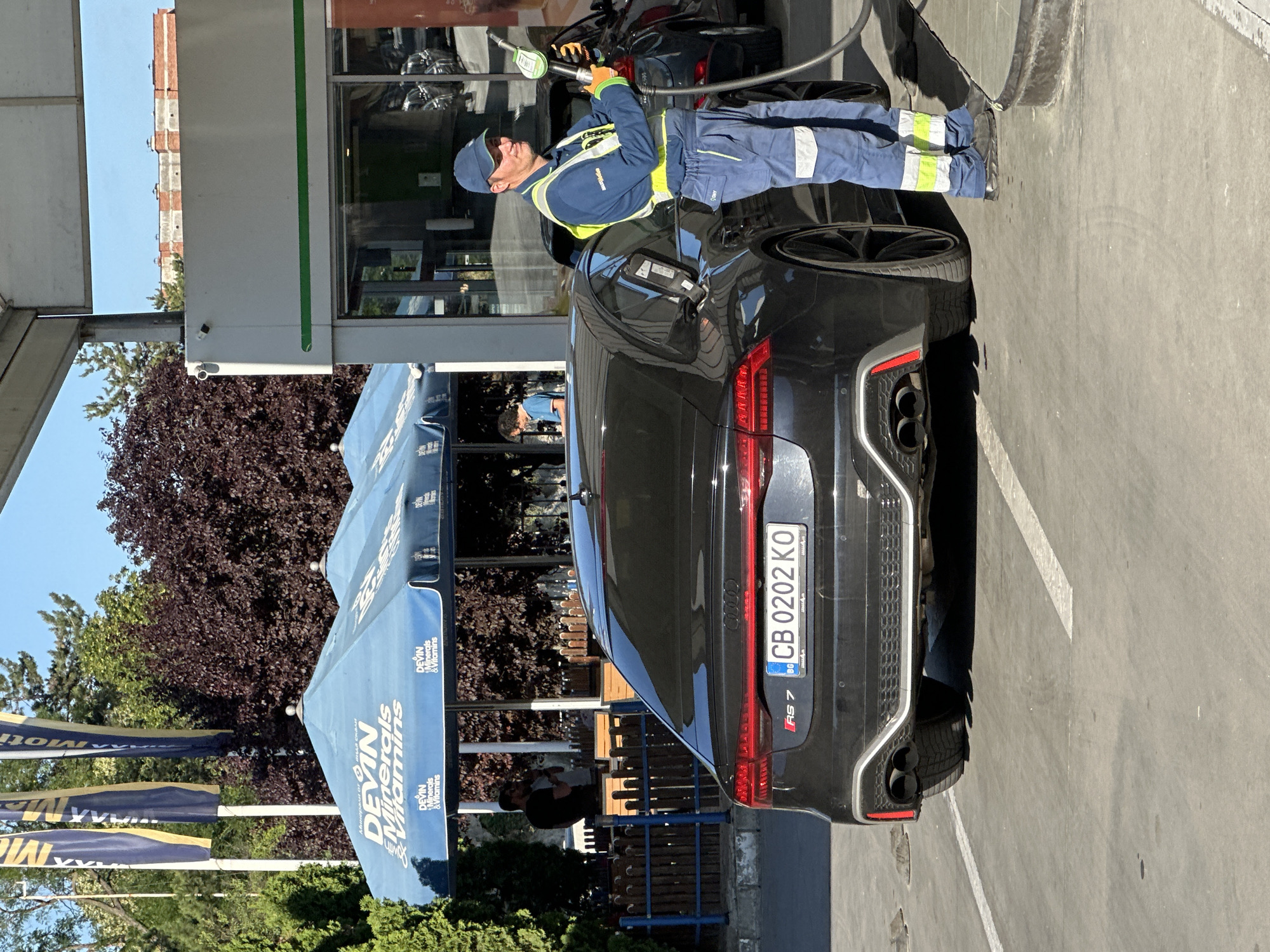 A gas station attendant in a blue uniform with reflective stripes and a blue cap is refueling a black Audi RS7 with a fuel nozzle. The car's license plate reads "CB 0202 KO". In the background, there are blue umbrellas with "DEVIN Minerals & Vitamins" branding, two men standing under them, trees, and a building. The scene appears to be outdoors on a sunny day. The location is the Lukoil gas station on Nikola Gabrovski Blvd in Veliko Tarnovo, Bulgaria.