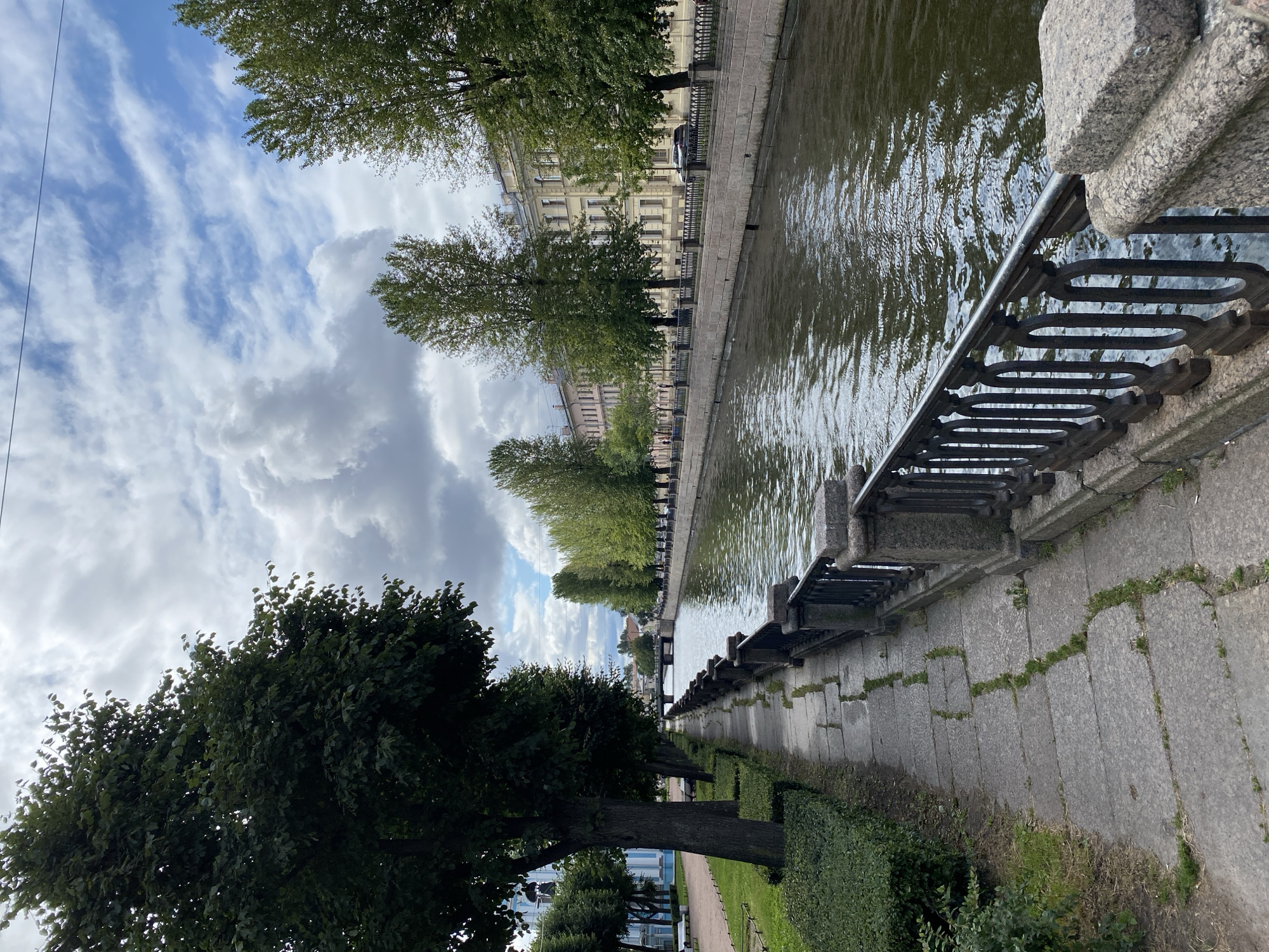 A picturesque canal lined with trees and buildings runs through a city. On the left, a paved walkway follows the canal, bordered by manicured bushes and large trees. The walkway is constructed of large, irregular stone slabs with grass growing in the cracks. A black metal railing with ornate scrollwork separates the walkway from the canal. On the right side of the canal, a row of cream-colored, multi-story buildings with numerous windows is visible, backed by more trees. The canal's water reflects the cloudy sky and surrounding greenery. A bridge is visible in the distance, spanning the canal. The sky is partly cloudy with a mix of white cumulus clouds and patches of blue. A few parked cars are visible along the street in front of the buildings on the right. The scene appears to be a pleasant day in Saint Petersburg, Russia, likely in a historic district, given the architecture and canal system.