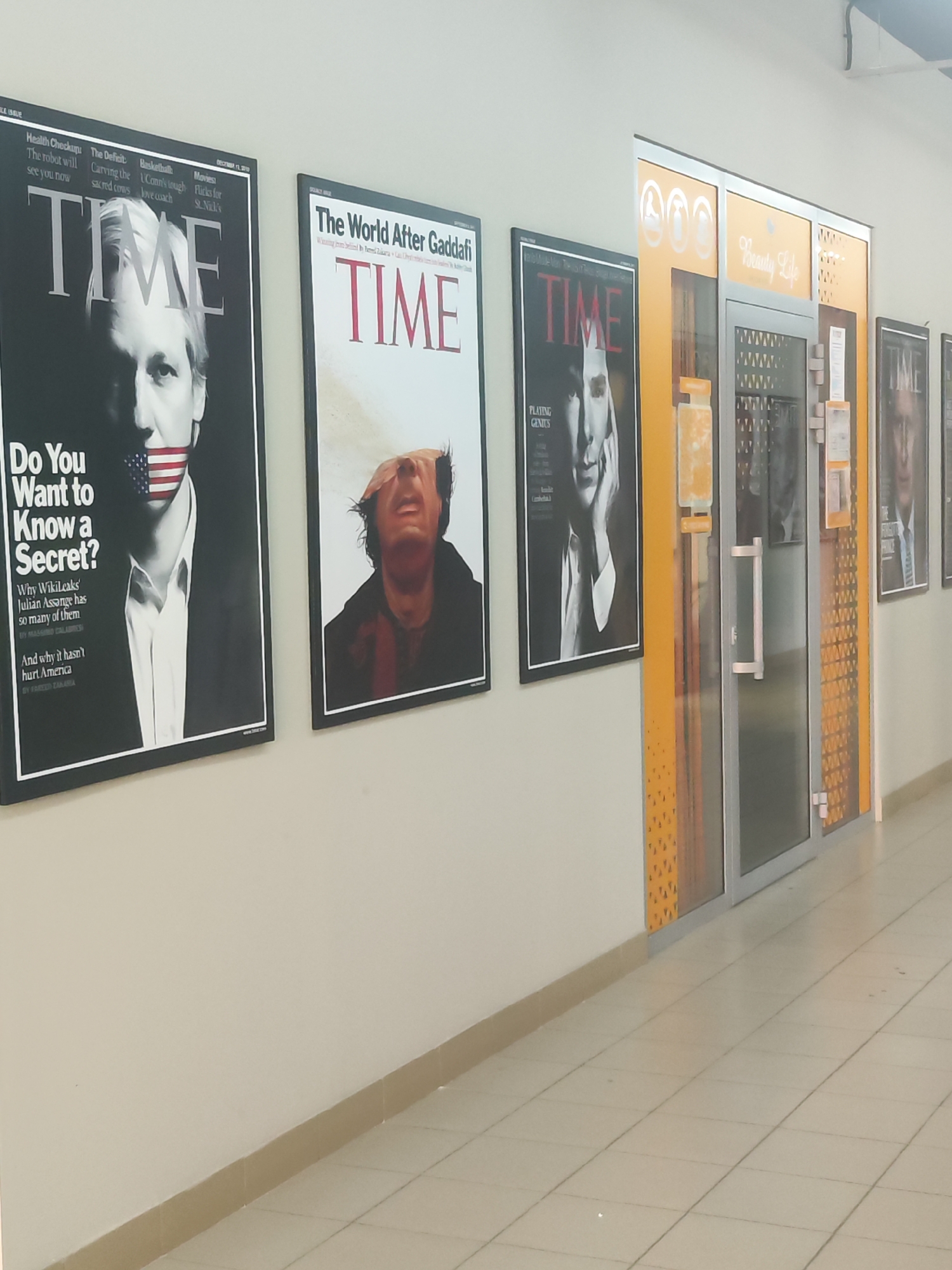 The image shows a hallway in what appears to be a building in the city of Chaikovsky, Russia. On the left wall, there are four TIME magazine covers framed and mounted. The first cover features a black and white image of a man with an American flag covering his mouth, under the title "Do You Want to Know a Secret?". The second cover reads "The World After Gaddafi." The third features a portrait with the title "Playing Genius." The fourth cover is partially visible and shows the forehead and eyes of a man. To the right, there is a doorway with the words "Beauty Life" printed above the entrance. The door is decorated with a design of orange shapes. The floor is tiled in a light color. The overall lighting is bright.