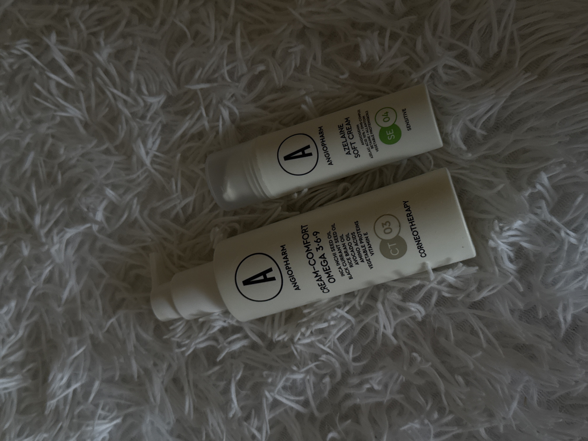 Two white bottles of Angiopharm skincare products are placed on a fluffy white surface, possibly a rug or blanket. The bottle on the left is taller and has a pump dispenser. It is labeled "CREAM-COMFORT OMEGA 3-6-9" and lists ingredients such as "INCA INCHI SEED OIL", "BLACK CURRANT SEED OIL", "RICE BRAN OIL", "AVOCADO OIL", "AMINO ACIDS", "VEGETABLE PROTEINS", and "VITAMIN E". It also has the designation "CT 03" and "CORNEOTHERAPY". The bottle on the right is shorter and has a screw-on cap. It is labeled "AZELAINE SOFT CREAM" and lists ingredients such as "ANGIOGENIN", "AZELAIC ACID 10%", "DMS-COMPLEX", "BETAINE", "ALLANTOIN", and "VEGETABLE PROTEINS/SOILS". It also has the designation "SE 04" and "SENSITIVE". Both bottles have a circular logo with the letter "A" inside. The lighting is dim, casting shadows on the fluffy surface. The scene does not contain any identifiable geographical markers indicating the city of Abinsk, Russia.