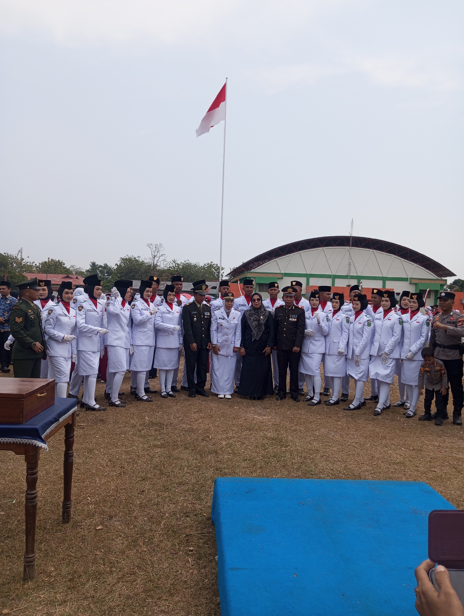 A group of people, mostly in uniform, stand in formation outdoors. In the foreground, a wooden table with a blue cloth is set to the left. Behind the table, a group of individuals dressed in white uniforms with red accents and black headwear stand in a line, facing the camera. To their right, men in dark uniforms with medals are positioned. Further to the right, a uniformed police officer stands, and a young boy in a police-like uniform is visible. A building with a curved roof is in the background, and a flagpole with the Indonesian flag is to the left of the building. The ground is covered in dry grass. The scene appears to be in Pematang Reba, Indonesia.
