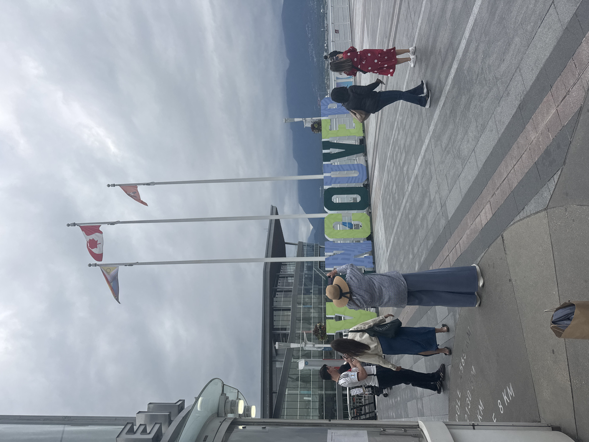 The image shows a group of people standing near the "VANCOUVER" sign in Canada Place, Vancouver, British Columbia. The large, colorful sign is made of block letters and is a popular tourist attraction. Three flags are visible above the sign: the Canadian flag, and two other flags, one with a yellow sun and red, yellow, and blue patterns, and another with a red maple leaf symbol. In the foreground, there are markings on the pavement indicating distances to various locations, such as "POLE 4565 KM" and "E 8 KM". Several people are visible, some taking photos of the sign. One person is wearing a red dress with white polka dots. The sky is overcast and cloudy, with mountains visible in the distance. The scene appears to be a public waterfront area with a modern building in the background.