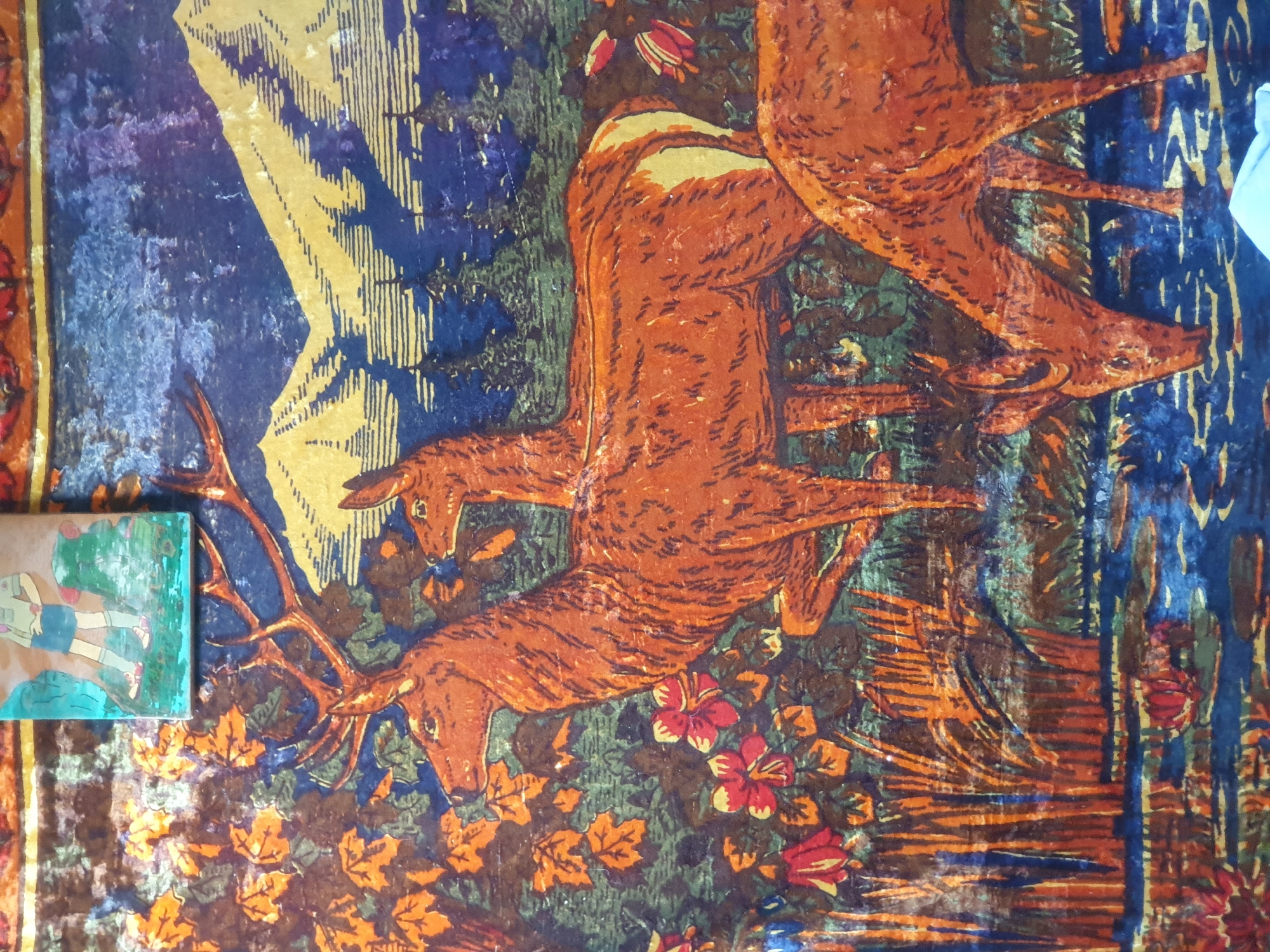 The image depicts a decorative panel featuring a naturalistic scene of deer in a woodland setting with mountains in the background. The primary subjects are three deer, rendered in warm, earthy tones. The largest deer, prominently positioned in the center, is a stag with large antlers. A second deer, smaller and appearing to be a doe, is partially obscured behind the stag. A third deer, with its head bowed to drink from a body of water, is visible in the foreground. The environment includes lush foliage with orange and red flowers, tall grasses, and a blue body of water with lily pads. In the distance, stylized green trees stand before a backdrop of yellow and blue mountains. The image appears to be a printed or painted artwork, possibly a tapestry or a decorative tin. The upper left corner of the image contains a small, separate image of a figure, possibly a boy in a green shirt and blue shorts. The artwork is not located in Zubrești, Moldova, but is a general depiction of nature.