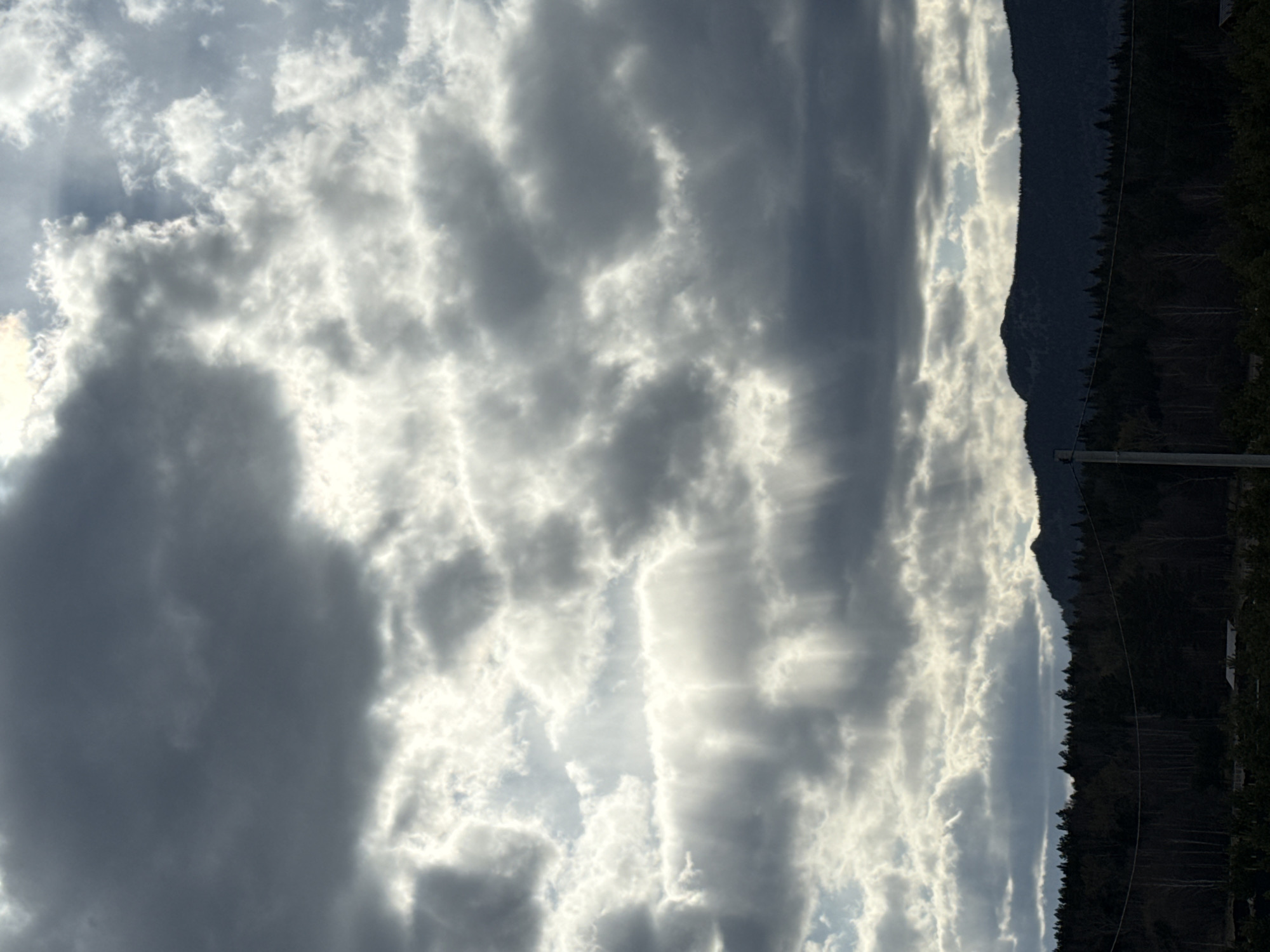 The image displays a dramatic mountainous landscape under a dynamic, cloudy sky. The sky occupies the majority of the frame, filled with dense, textured clouds ranging from brilliant white and glowing where sunlight penetrates, to various shades of dark gray in shadowed areas. Visible sunbeams, or crepuscular rays, break through the clouds, especially towards the upper left, adding depth and a sense of atmosphere.
Below the expansive sky, a prominent mountain range forms a dark, rugged silhouette across the lower portion of the image. The mountain slopes are covered with a dense forest, appearing as an unbroken dark mass that outlines the mountain's contours. The sharp contrast between the bright, illuminated clouds and the dark, shadowed mountains creates a striking visual effect.
A utility pole and faint power lines are visible in the lower right quarter, suggesting some human presence or infrastructure in the otherwise natural and wild setting. No people or specific activities are discernible. The scene depicts a powerful moment in nature, likely during daytime with variable weather, characteristic of a mountainous region like Tyulyuk, Russia.