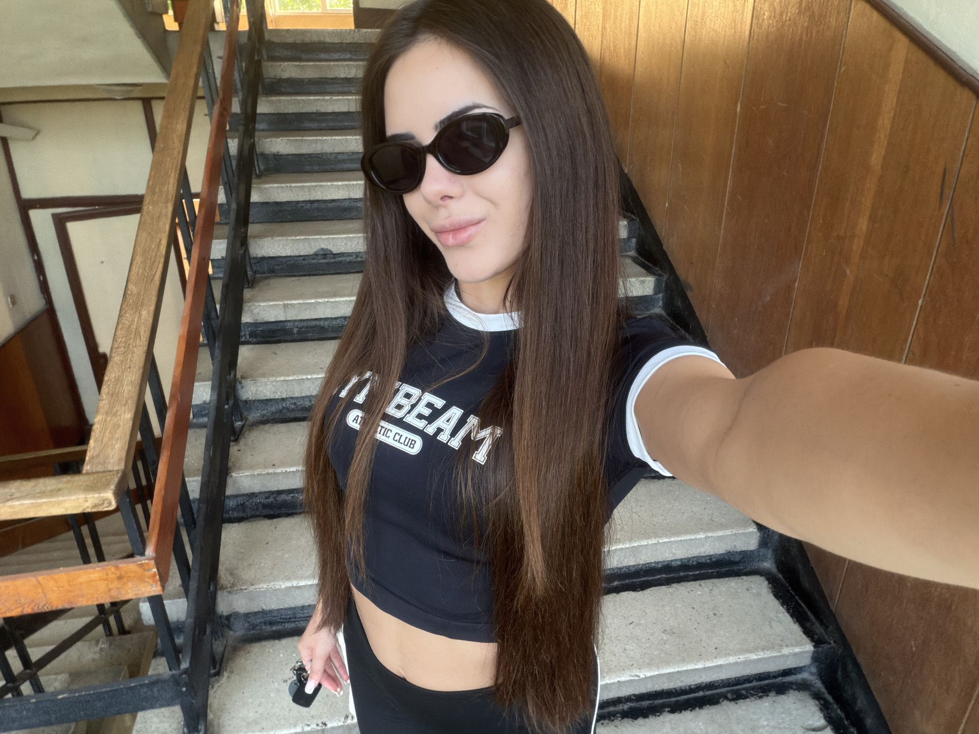 A brunette woman stands on a concrete stairway, apparently taking a selfie. She wears sunglasses, a black crop top with white trim and lettering, and black leggings. The stairway has a wooden railing on one side and a wall on the other. The stairway appears to be in a building. The location is Sofia, Bulgaria.